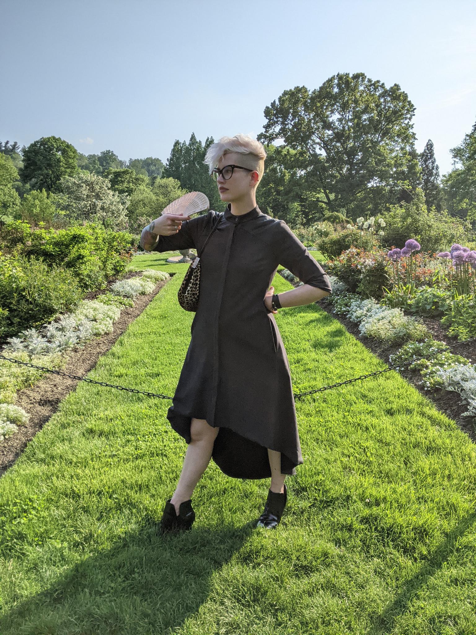 Nicole, a white genderqueer human, stands in a well manicured botanic garden in a dress, in a hot mean pose. It is also literally 99 degrees where they are. they have shaved sides and bleached hair.