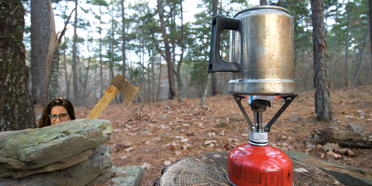 in the foreground, a kettle of water heats atop a camp stove balanced on a tree stump, in the background, behind a pile of rocks, kayla waits with a smile in her eyes, just peering above the rocks, a rusty axe leans out from behind the rock to the side. the whole thing is photoshopped and a little goofy.