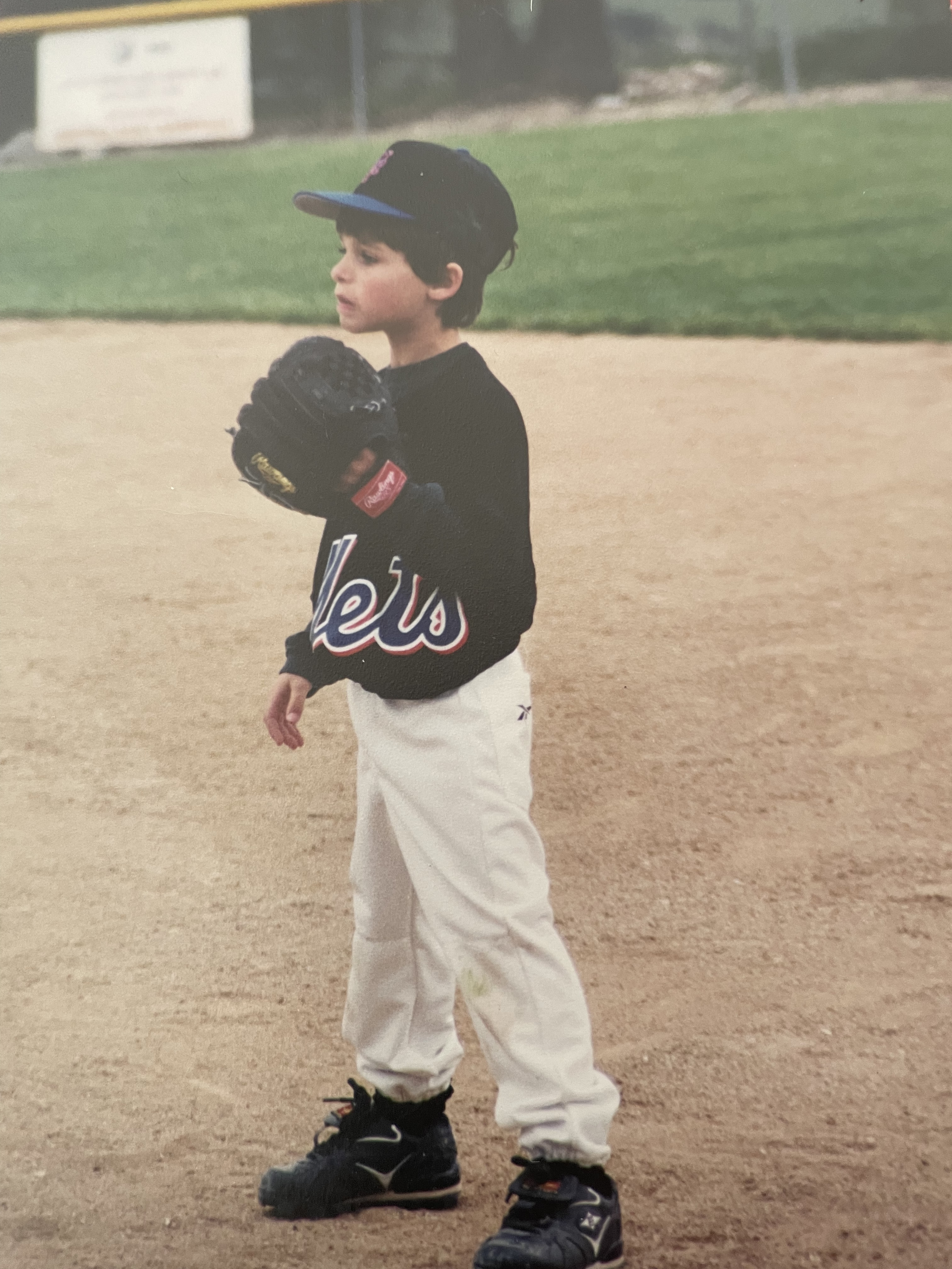 The writer, Drew, as a young child wears a Mets jersey, baseball hat, and baseball glove while standing in a baseball diamond.