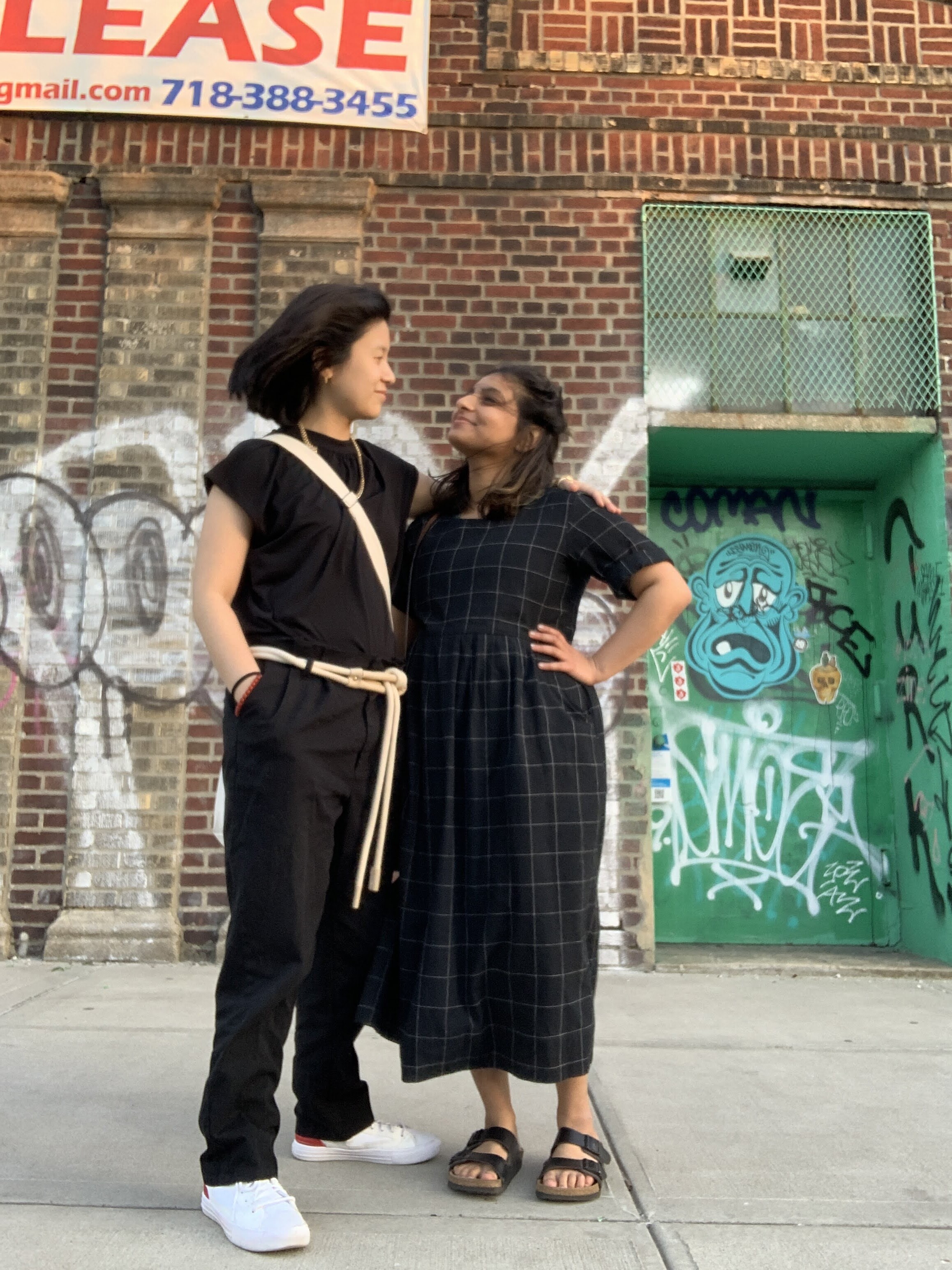 Gerrie and Ashni post in this photo in front of a graffitied brick building. Gerrie is wearing all black with a rope-like belt and white sneakers. Ashni is wearing a long dark grid-patterned dress and birkenstocks. They are looking at each other romantically or happily