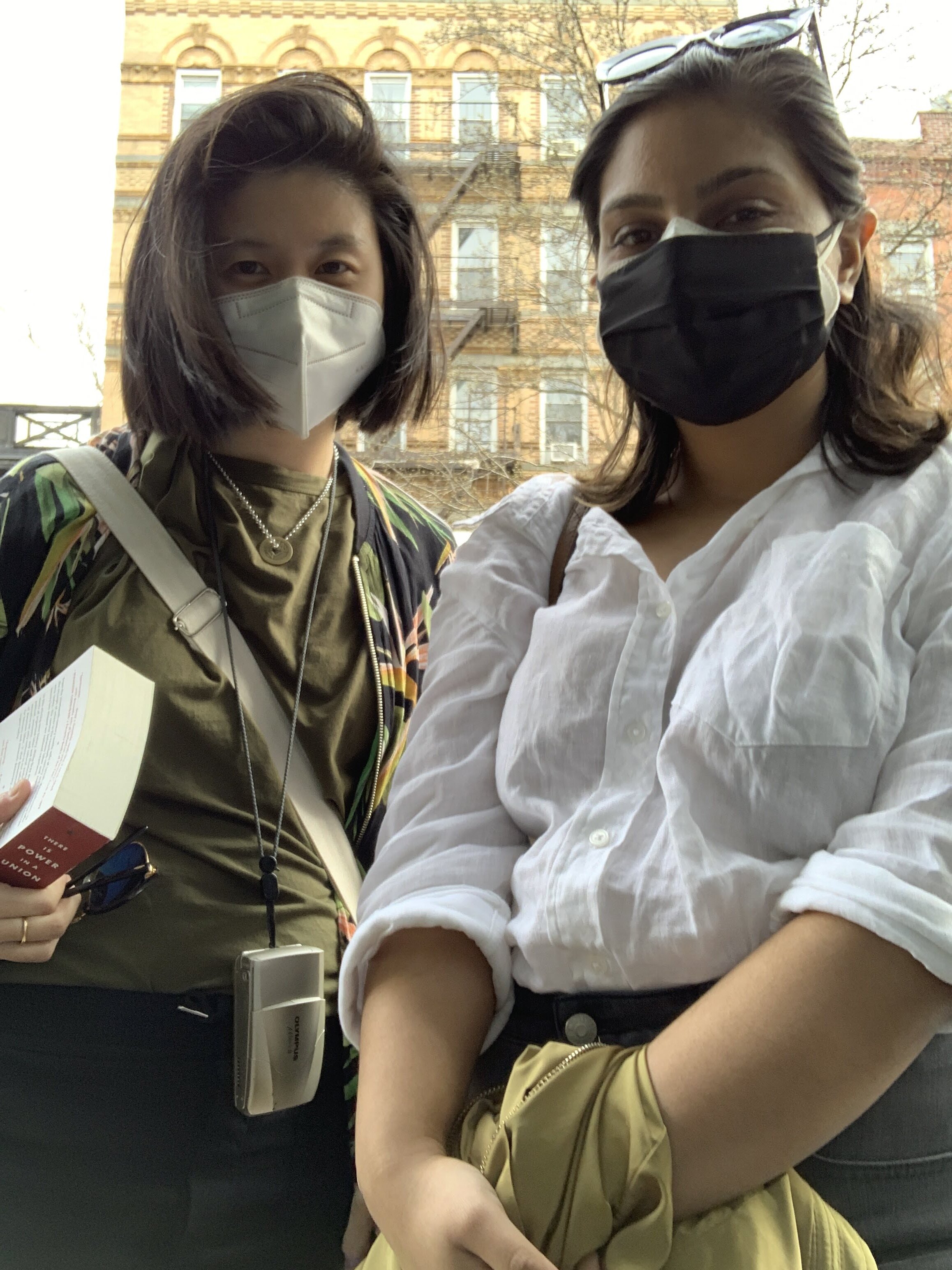 Gerrie and Ashni pose for a photo while wearing masks. They are outdoors and look like they are on their way somewhere with bags on their shoulders and such