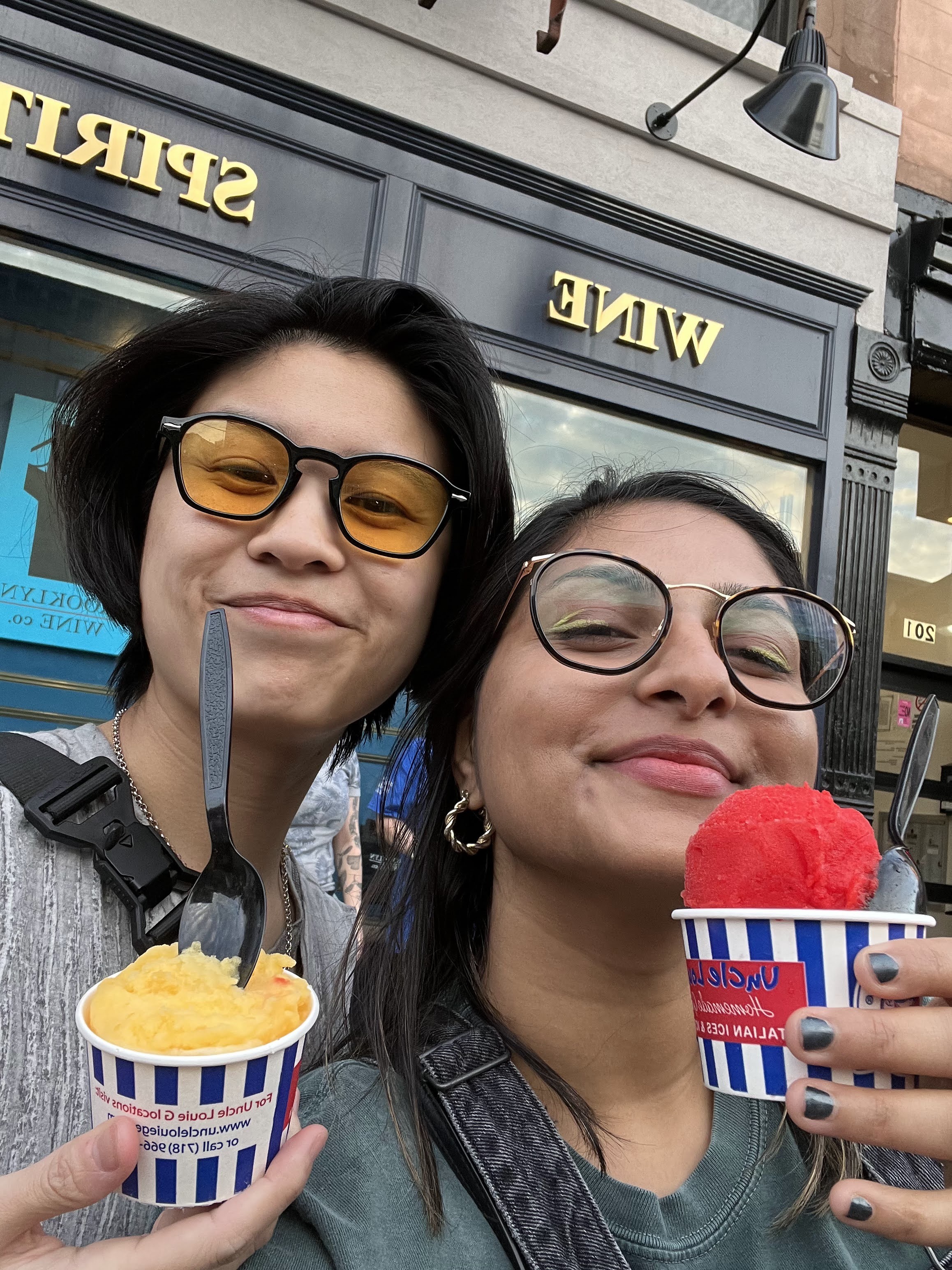 Gerrie, a Chinese-Filipino nonbinary person has a short haircut and black hair and is wearing yellow tinted sunglasses and smiling. Next to them, Ashni, a gender averse South Asian person wearing prescription glasses is also smiling. They are both holding up italian ices, one yellow, one red.