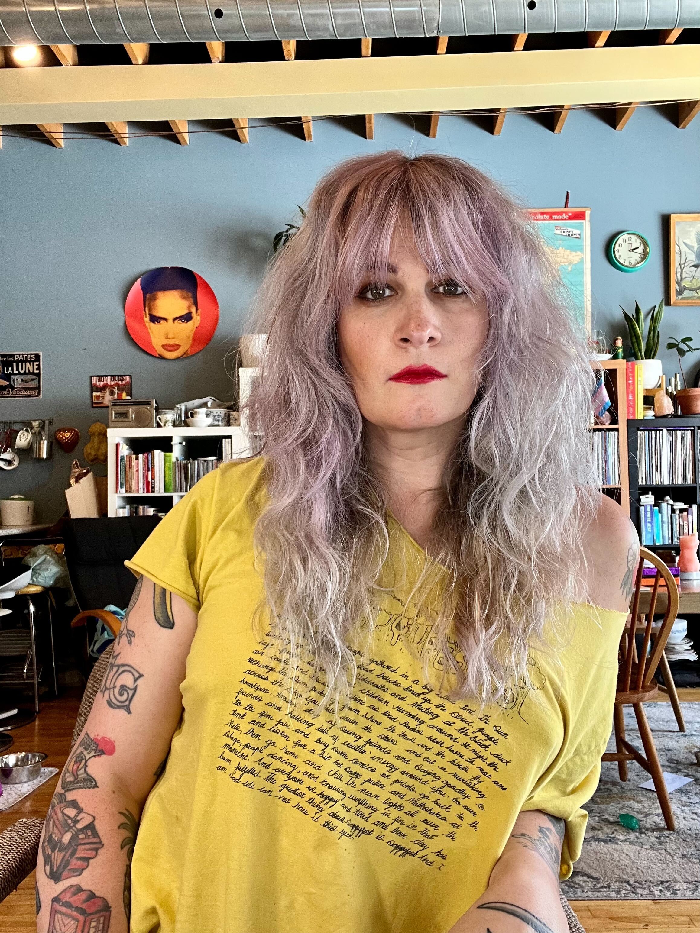 Niko is a white woman pictured here wearing an off the shoulder yellow tee shirt and bright red lipstick, while in her eclectically and enviably decorated apartment. She has tattoos and a platinum blonde to lavender-ish blow out hair style with bangs and long waves. 