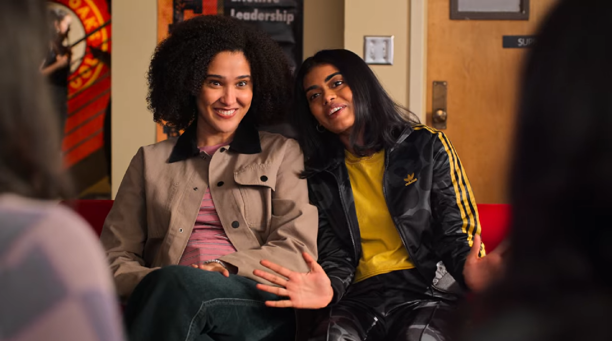 Fabiola and Aneesa sit with their shoulders pressed, smiling. Aneesa leans into Fabiola with her hands outstretched.