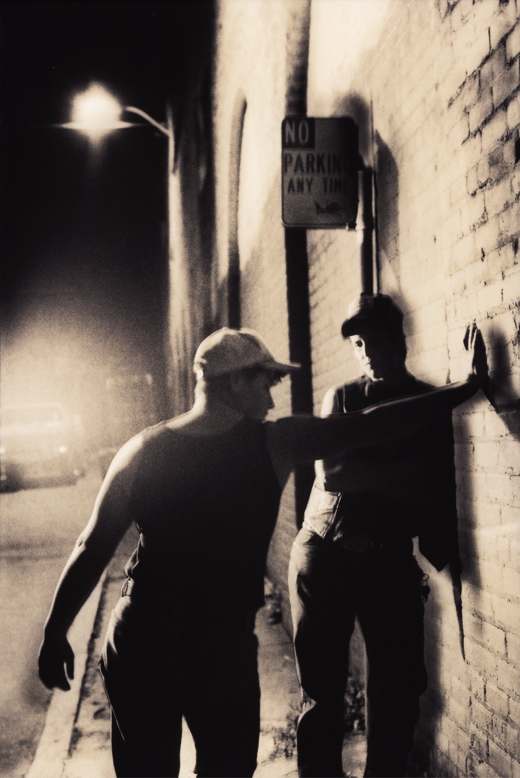  A black and white image shows the silhouette of a masculine person wearing a baseball cap and jacket leaning against a brick wall in front of a "No Parking Any Time" sign. Another masculine person, whose back is to the camera, stretches out their arm to press their right hand against the brick wall. They wear a baseball cap and a black tank top. A street lamp is visible in the background.