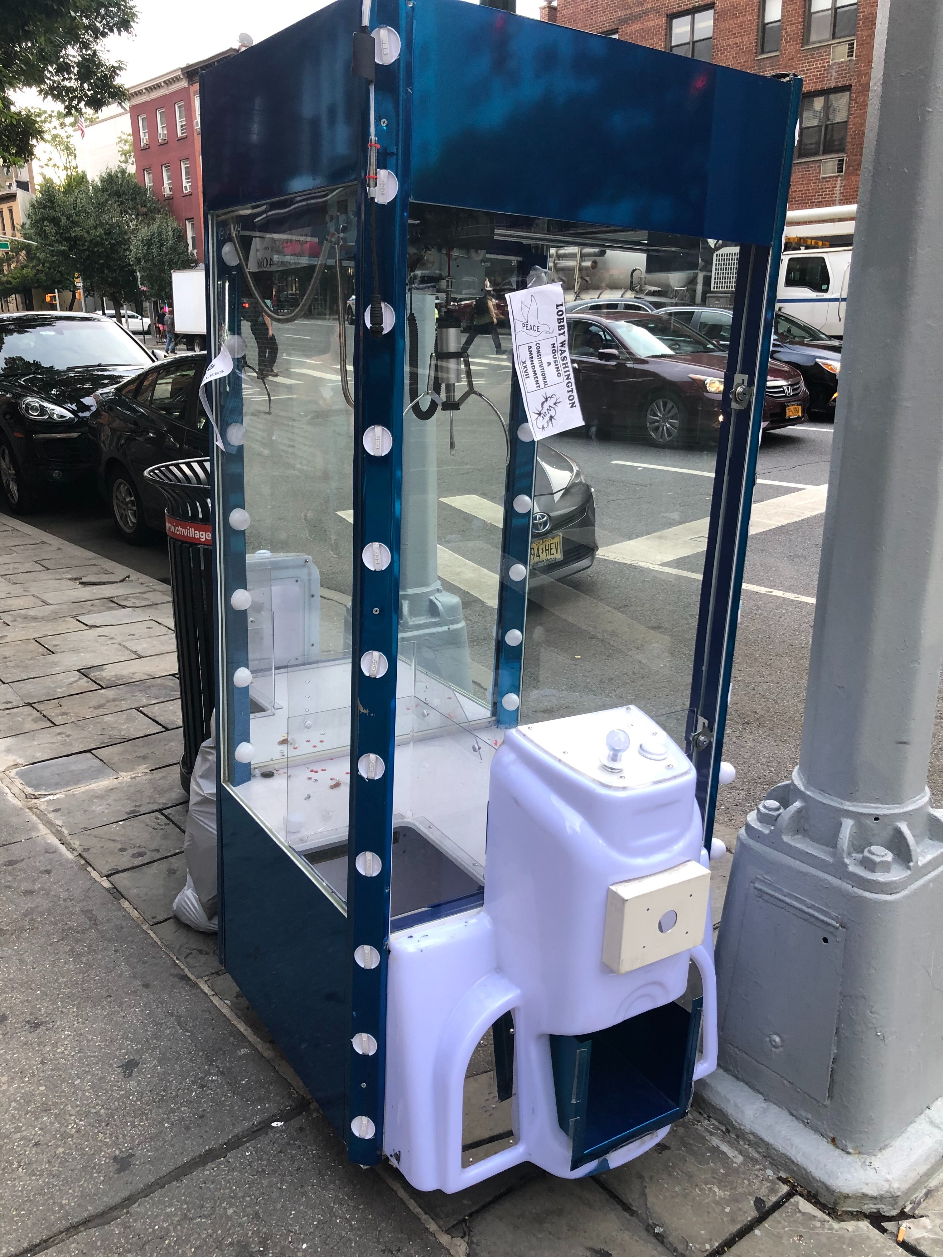 a large, empty blue claw machine lies abandoned on a street in new york city, it's lights unlit, its chamber, lacking prizes