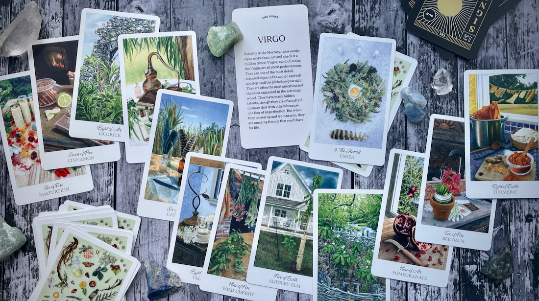 A collection of various tarot cards are strewn on a grey wood tabletop surface, shot from above. In the center is a card with the word "Virgo" in large print and small print description text that is not visible. There are also various crystals.