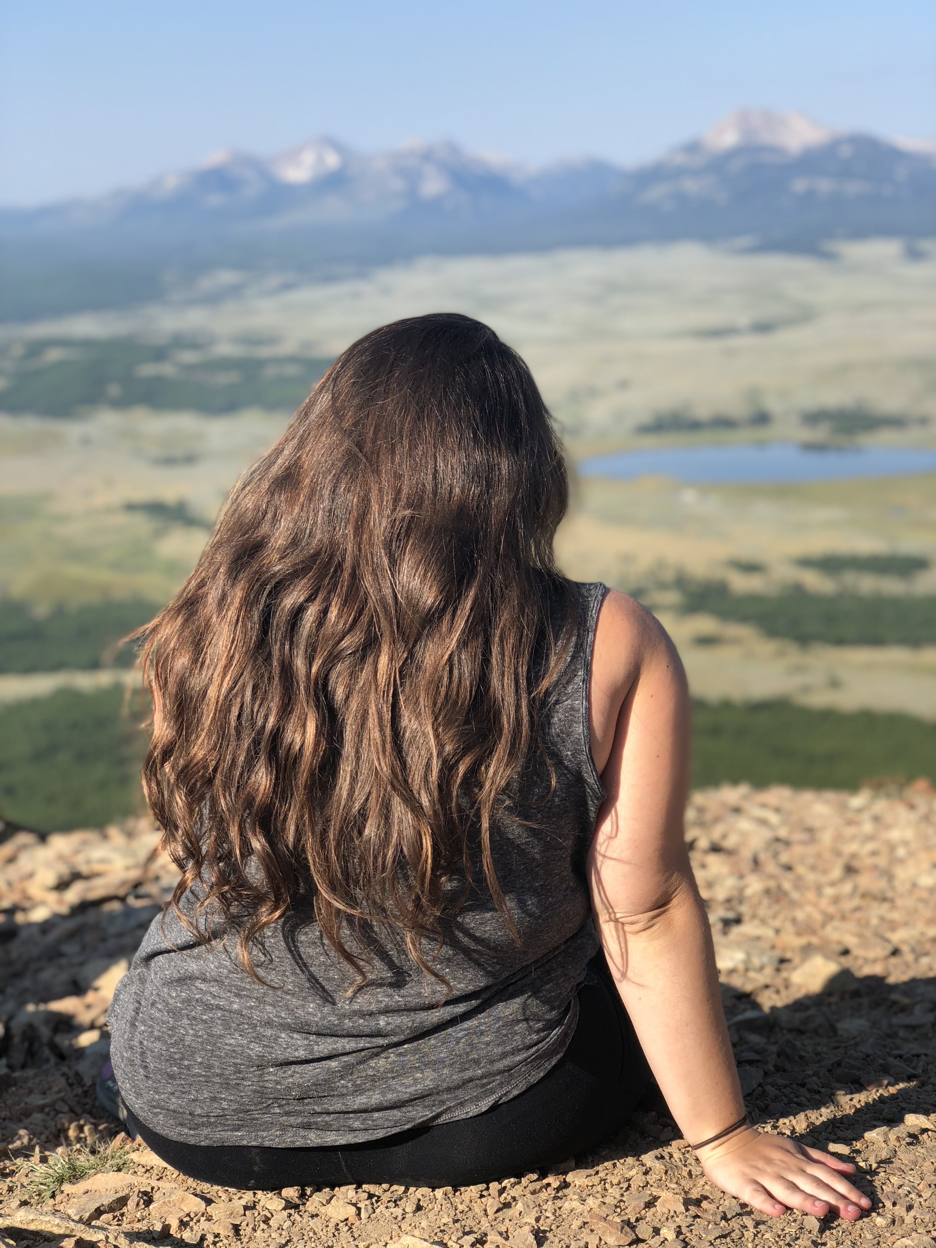 Vanessa sits on a hill overlooking a beautiful natural landscape with mountains, a lake, and fields. She has long brown hair and is a white woman.