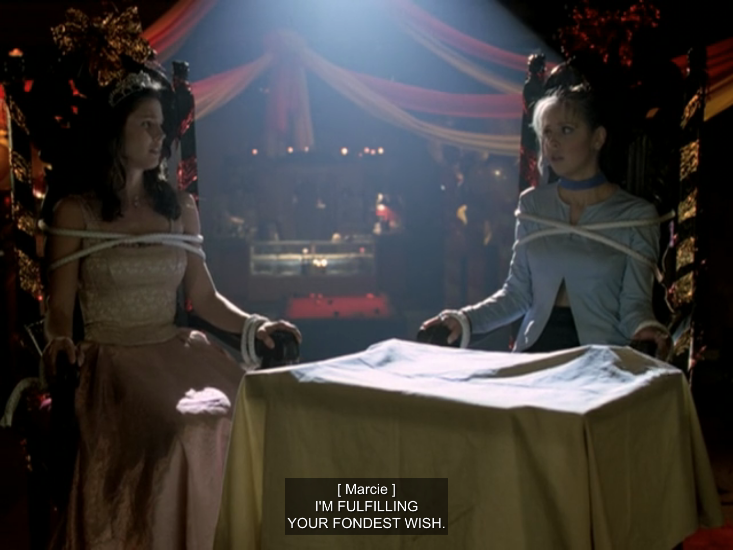 Buffy and Cordelia tied up in chairs in a dark hall that’s ready for the Homecoming dance. They’re looking at one another in horror. Off screen, Marie is saying “I’m fulfilling your fondest wish.