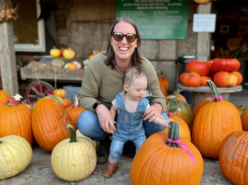 Casey a white woman with shoulder length brown hair poses with her small child in a pumpkin patch