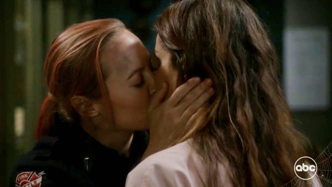 Maya, who has pink hair pulled into a ponytail, kisses Carina at the firehouse on Station 19