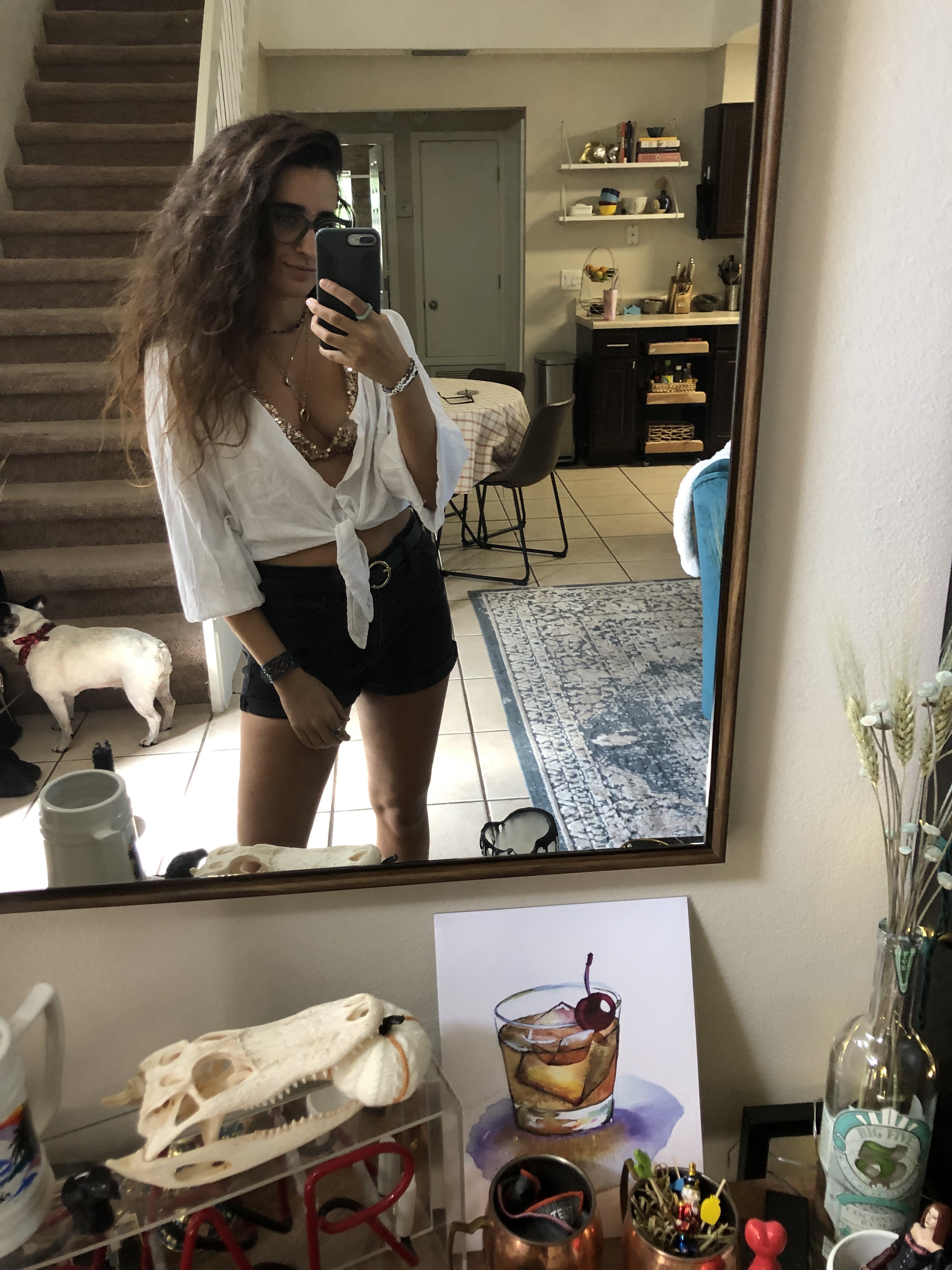 Kayla Kumari Upadhyaya wears black denim shorts, a belt, a tie-front white shirt, a gold bikini top, and blue framed glasses. Her long curly brown hair is down.