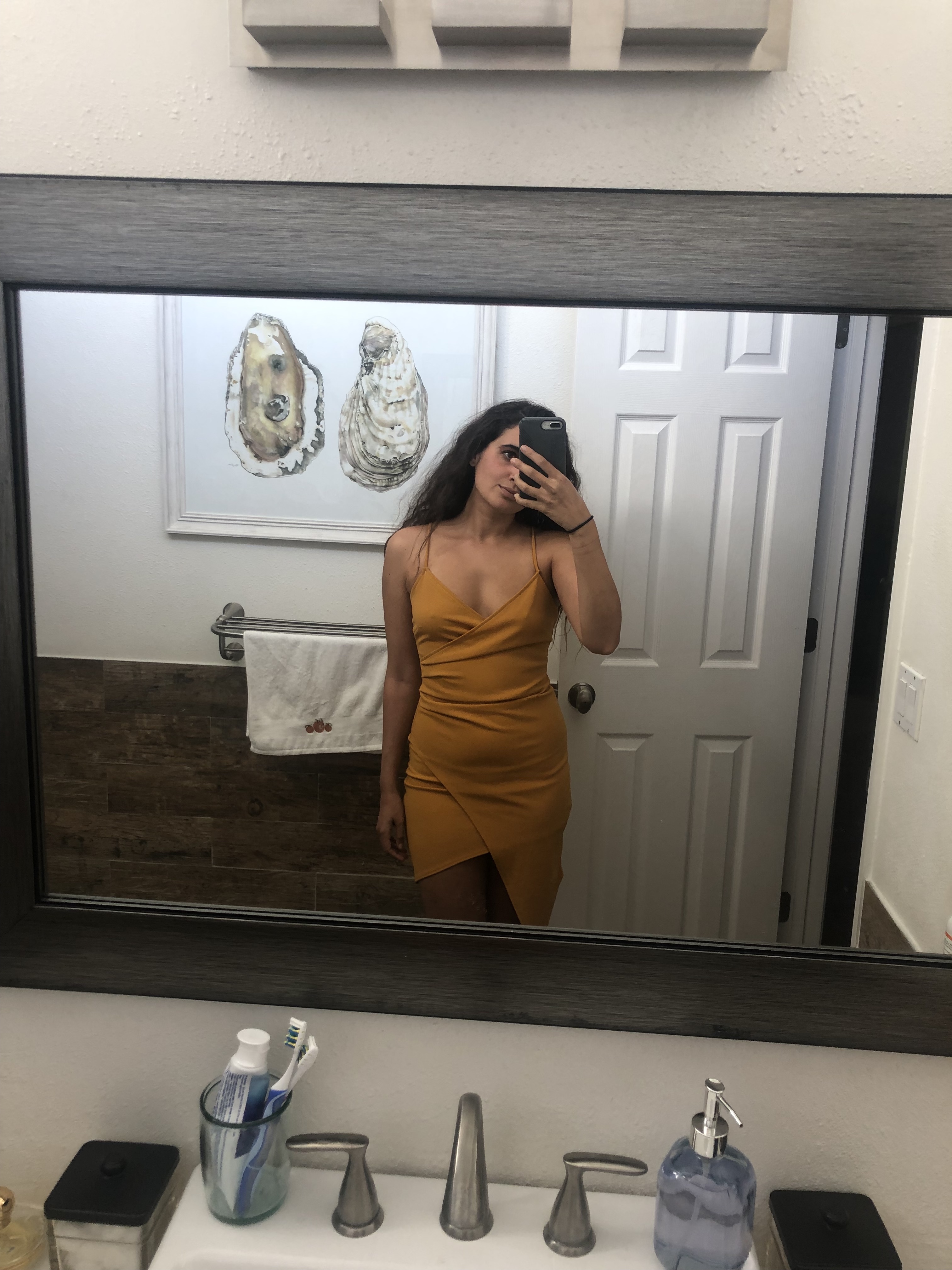 Kayla Kumari Upadhyaya wears an orange tight asymmetrical mini dress, and her long curly brown hair is down. An oyster painting is behind her.