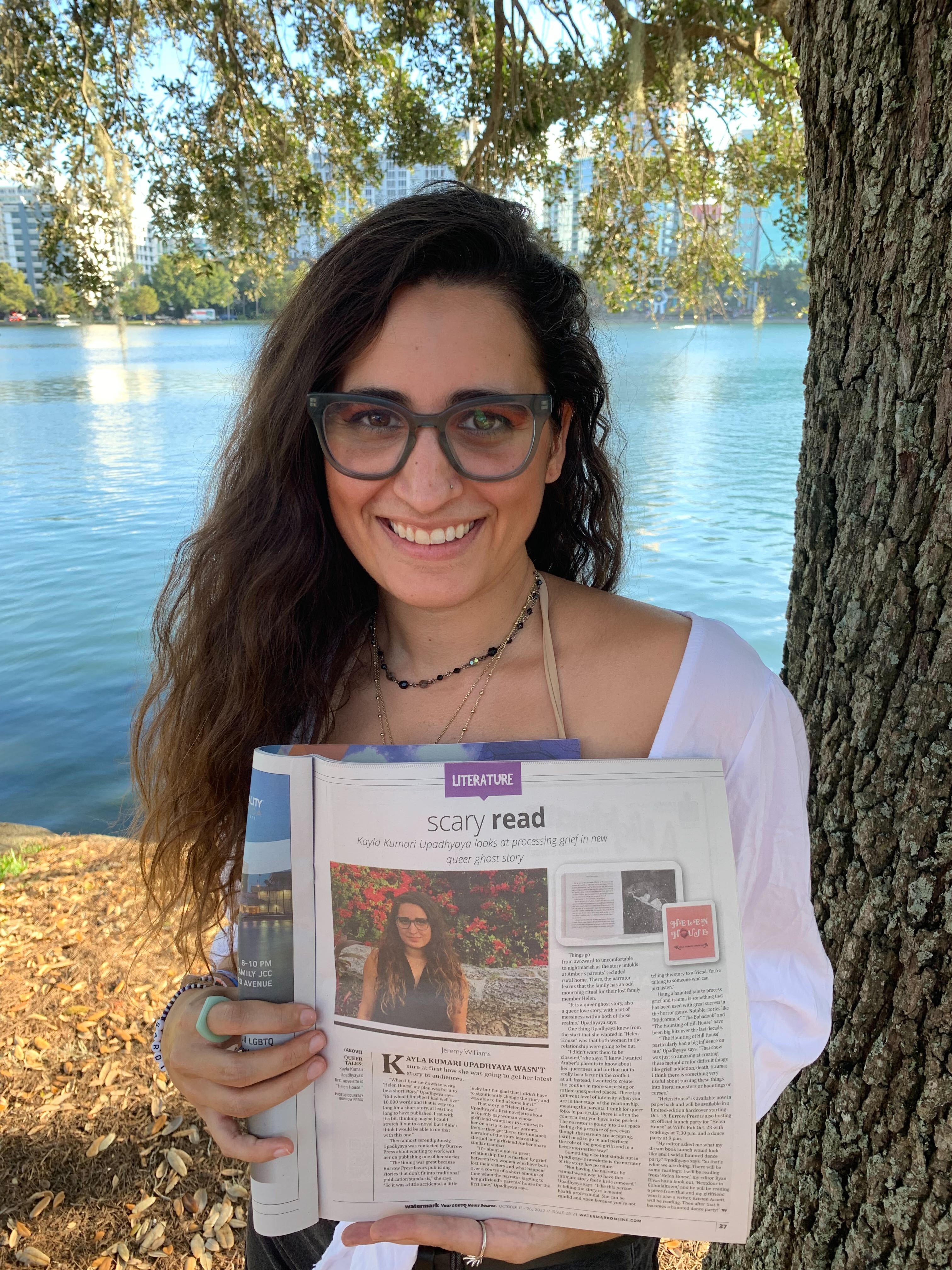 Kayla Kumari Upadhyaya stands against a tree near a lake holding up a newspaper article featuring her book Helen House