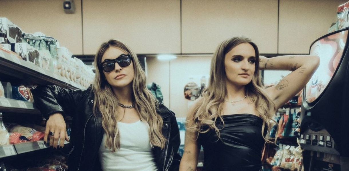 Chrishell Stause and G Flip are in a convince store dressed as each other, both are white people with shoulder length blonde hair, one has on a white tank top and black leather bomber jacket with large black sunglasses, the other is in a black sleeveless mini dress