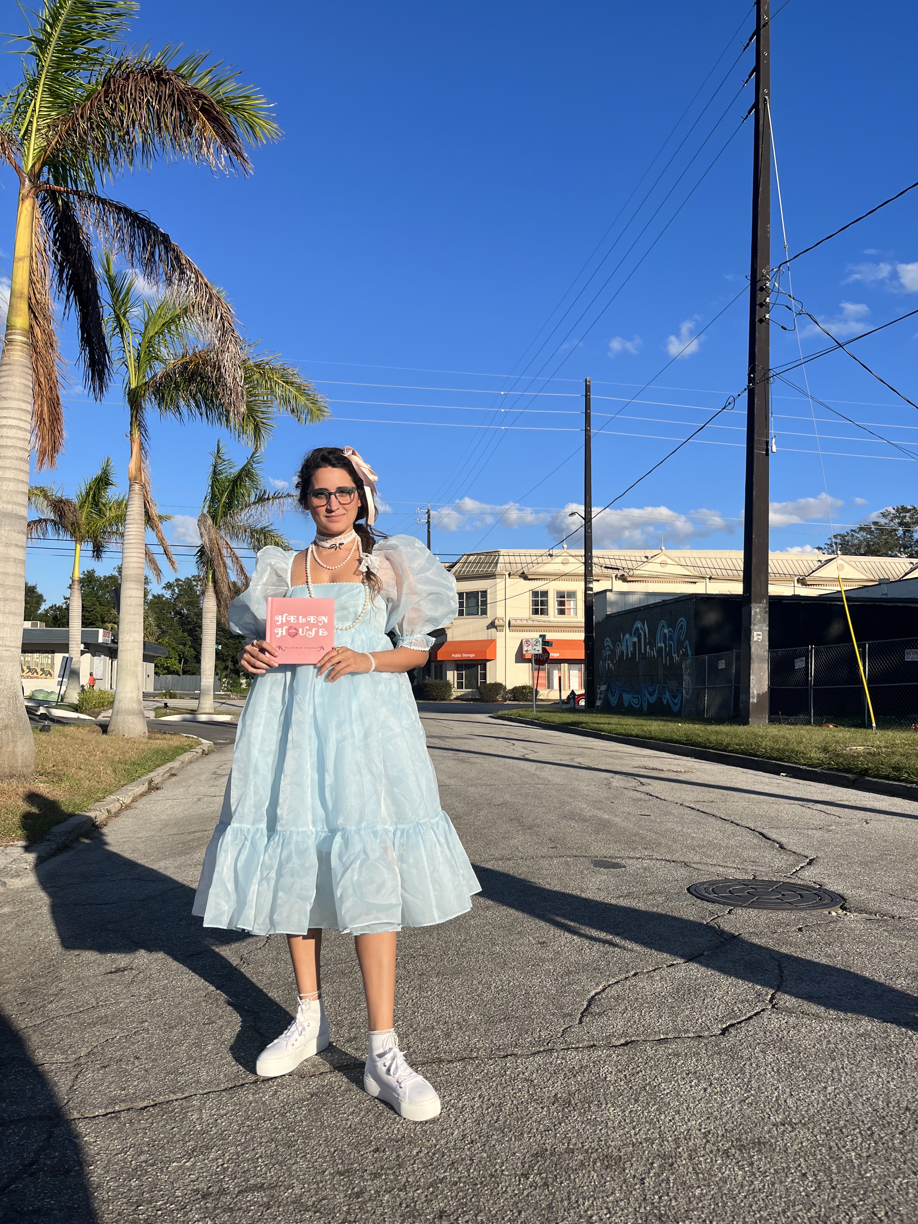 Kayla, a South Asian woman stands in a blue doll dress, holding her book, Helen House.