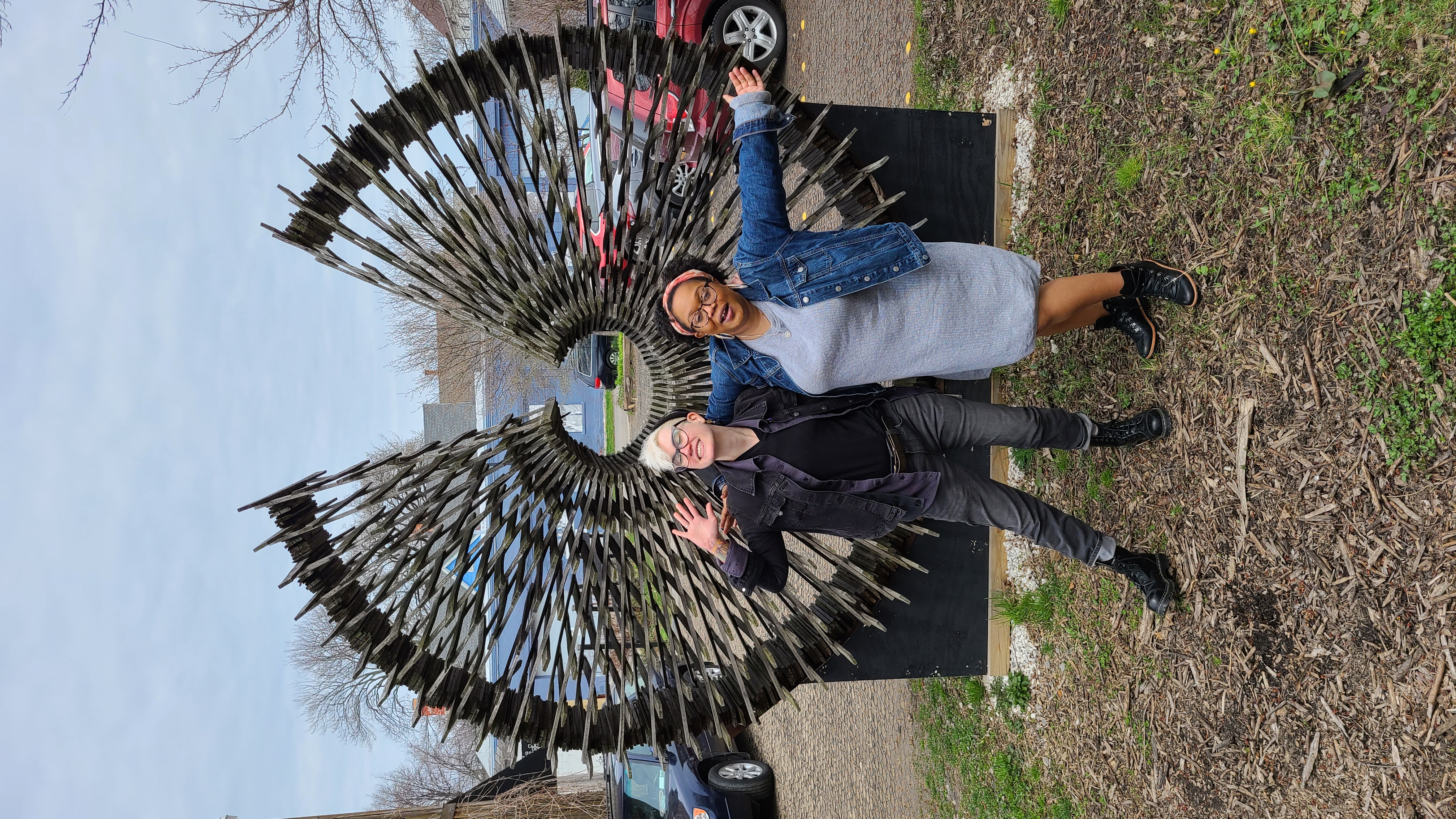 Carmen and Nico are hugging with their bodies open to the camera, they are in front of a metal sculpture that has an open circle with straight lines coming out. Nico is a white genderqueer person with blonde hair, a black denim coat, black shirt, and black jeans. They have on a black beanie. Carmen has on a pink headband, denim jacket, and grey sweater dress, and combat boots.