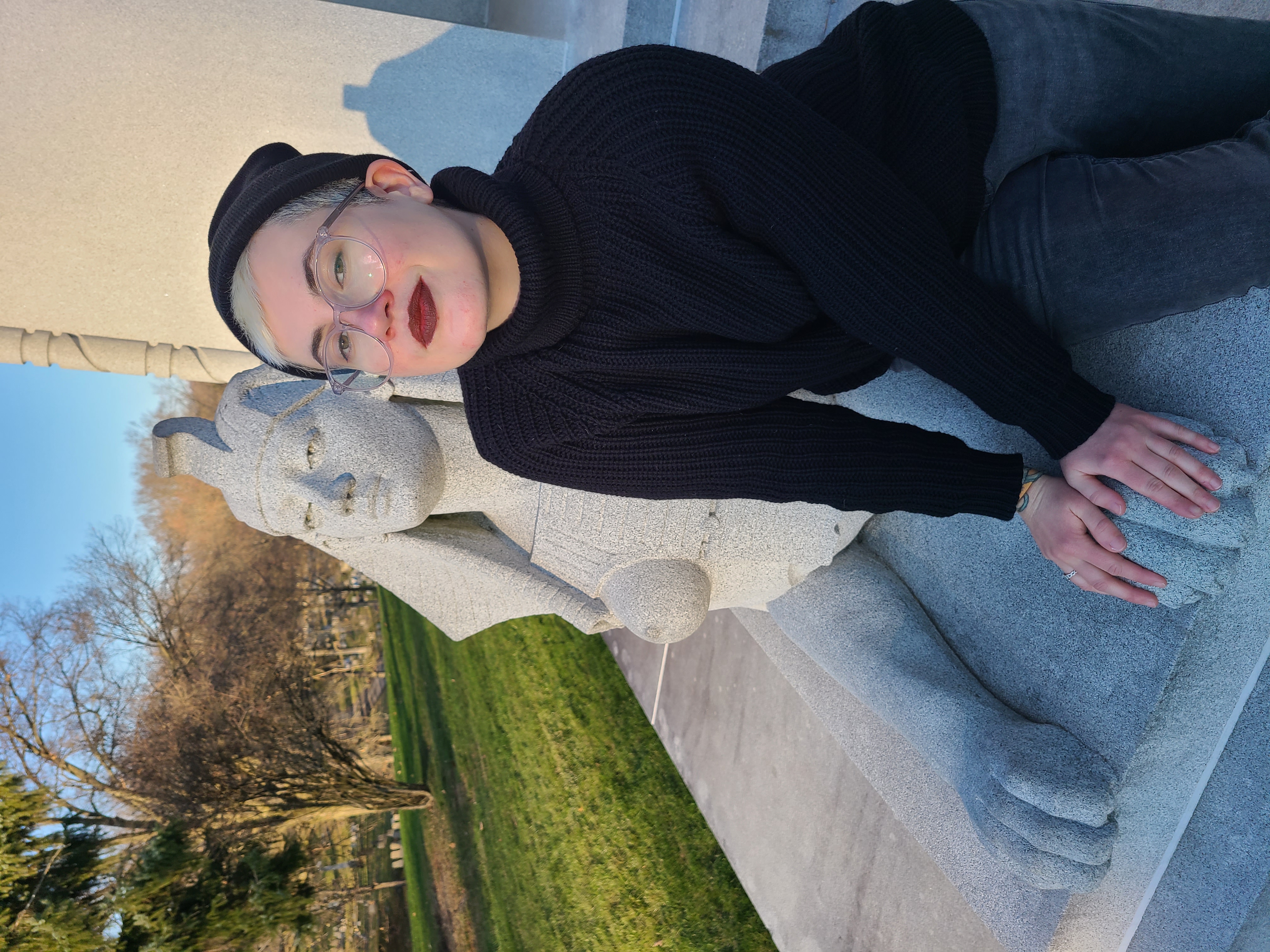 Nico, a white genderqueer human with blonde hair leans against a sphynx, wearing a black turtleneck and beanie as well as glasses and burgundy lipstick