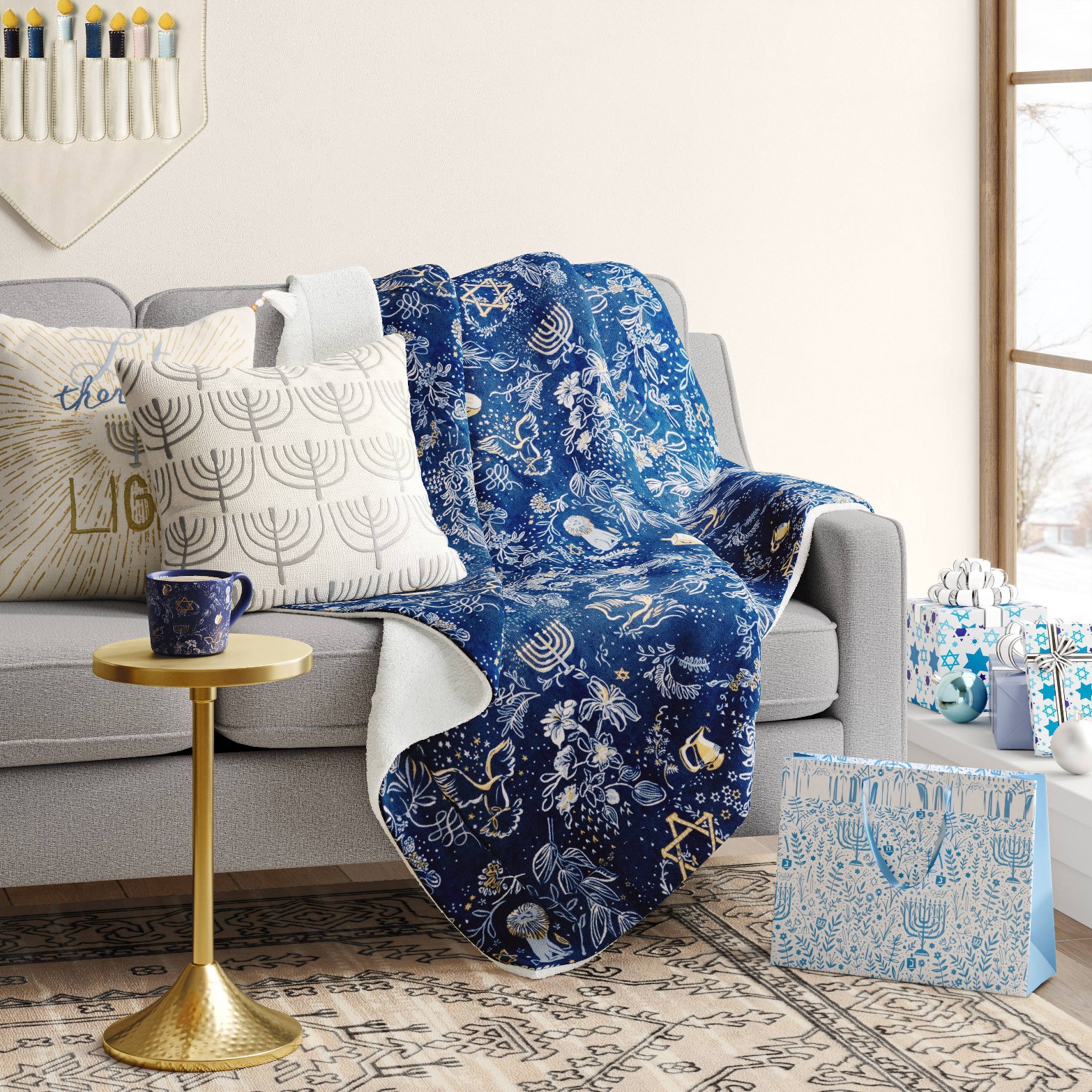 a blue blanket with gold hanukkah print: menorahs, star of david draped over a gray couch. there are gray throw pillows and hanukkah print gift bags on the floor.