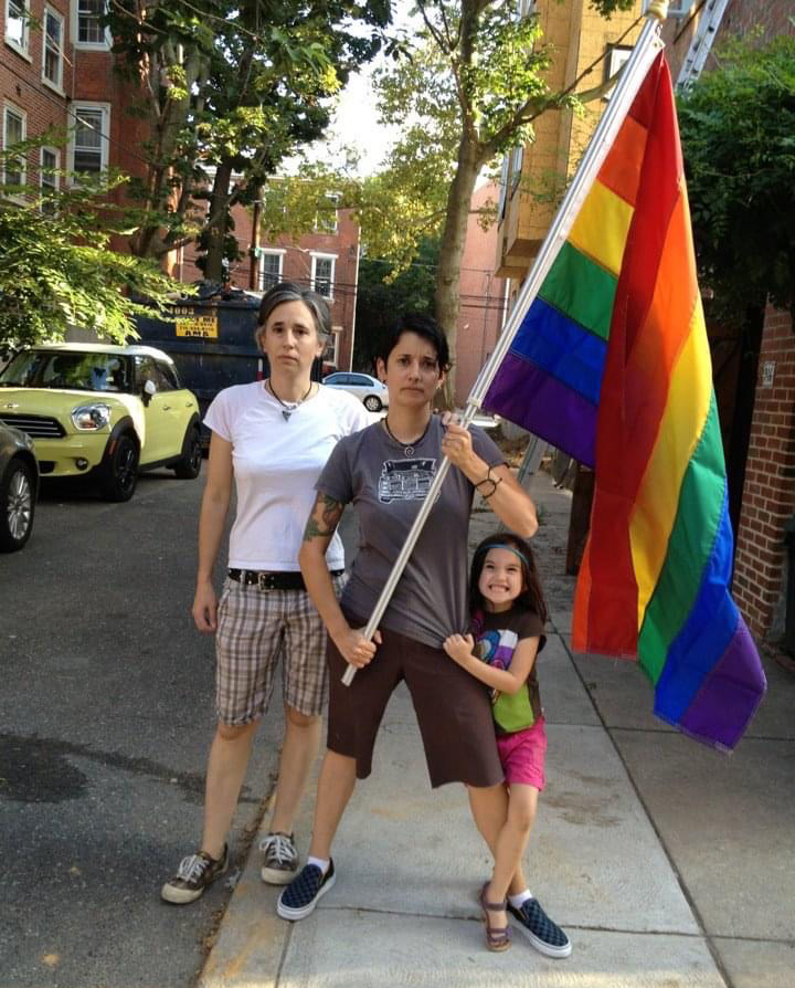 Tracy, Mia and their kid stand with a Pride flag. Tracy is holding the flag, her feet spread apart, looking very serious. Behind her Mia stands, straight and staring forward with a similarly serious expression. The kid grabs onto Tracy's shirt and smiles wildly.