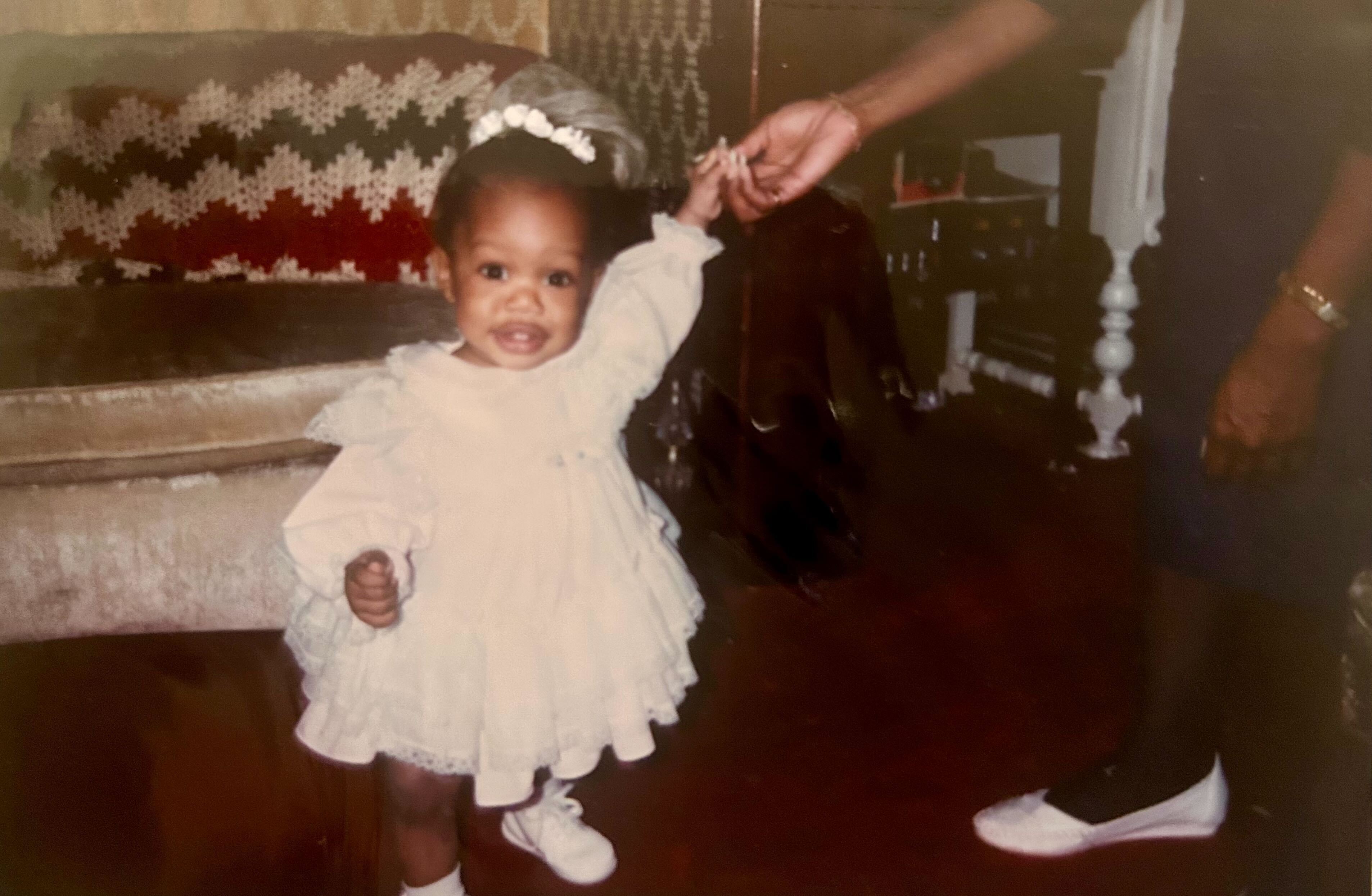 Carmen is at her first birthday party in a fluffy white dress and white flower in her hair, in white booties.