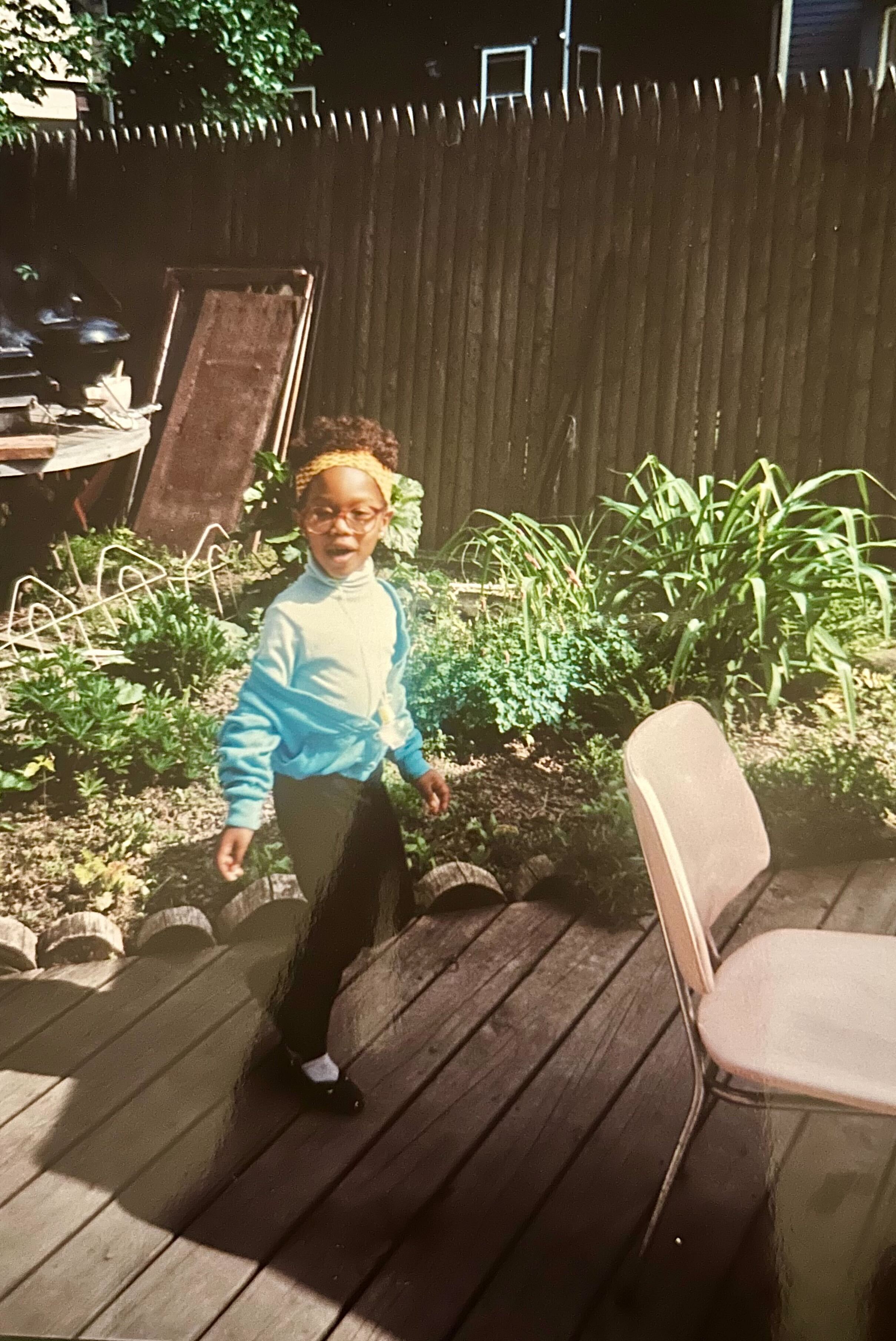 Carmen is about six years old and in a yellow headband, red glasses, and turquoise blue sweater set with black leggings, she's running around her Aunt Lorna's backyard.