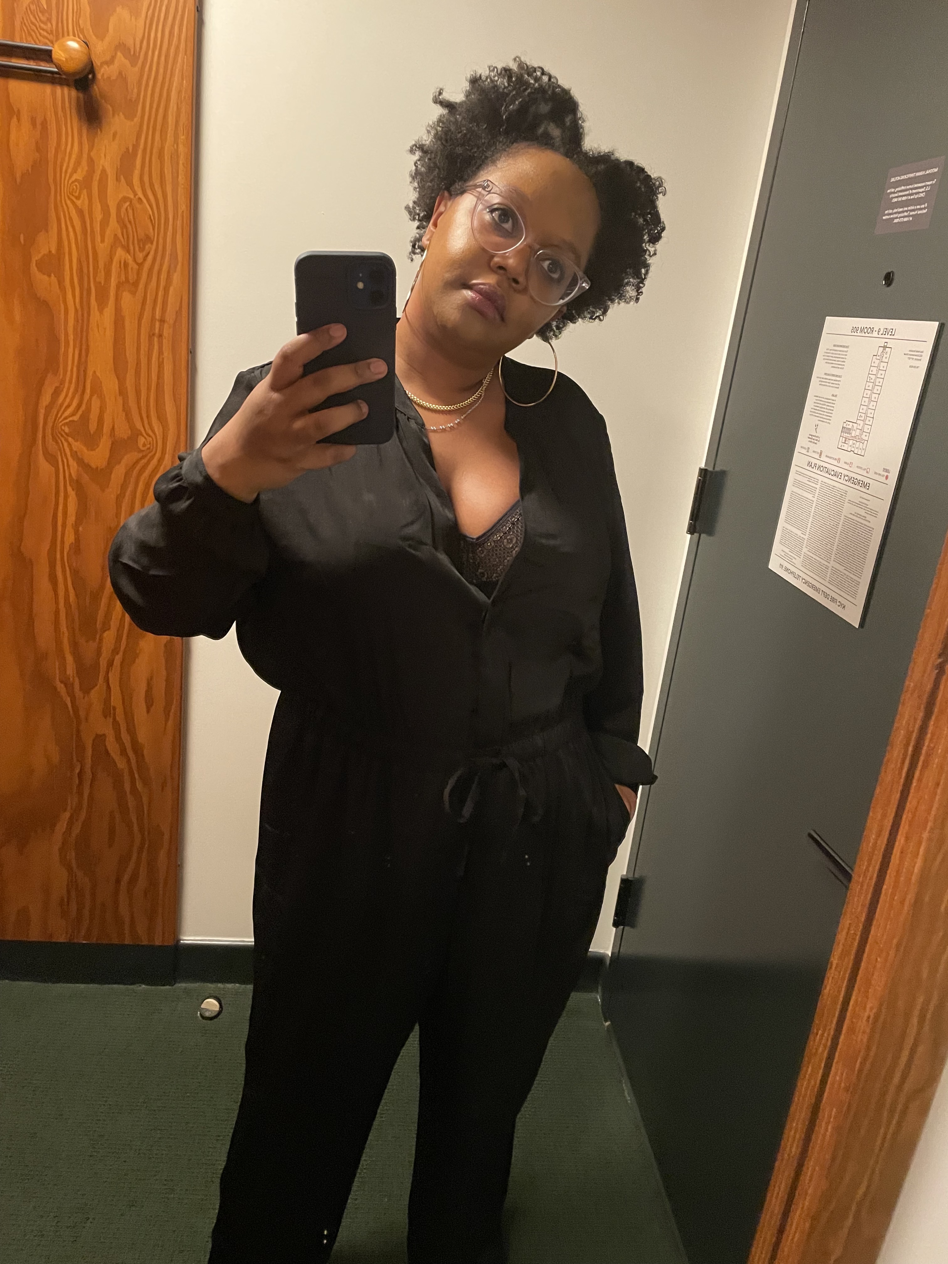 Carmen is in a black silk jumper that is unbuttoned to show a little black lace bra and has large gold hoop earrings and a gold chain necklace. She's in a hotel mirror, taking a selfie.