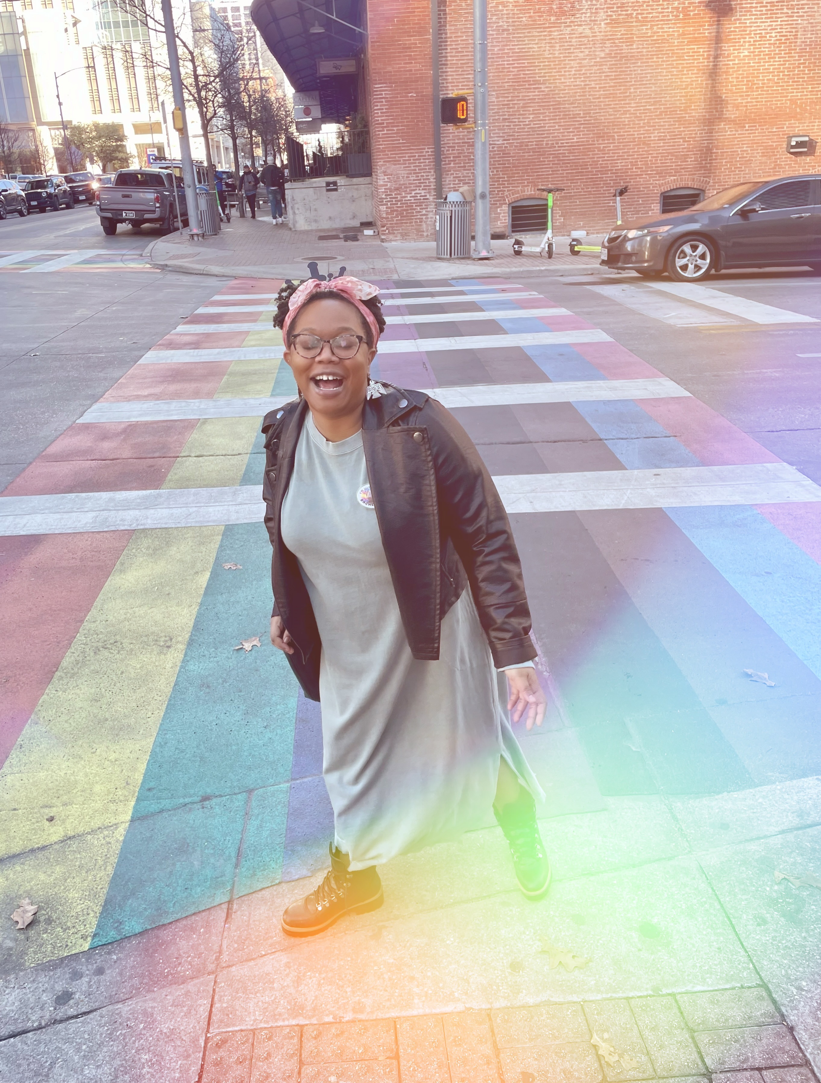Carmen, a Black femme, is in a green long dress with a Black leather motorcycle jacket and her hair bulled up in a pink scarf. She has on combat boots. She's standing outside on a street corner with where the street is painted rainbow and also there is a rainbow sun glare on the camera.