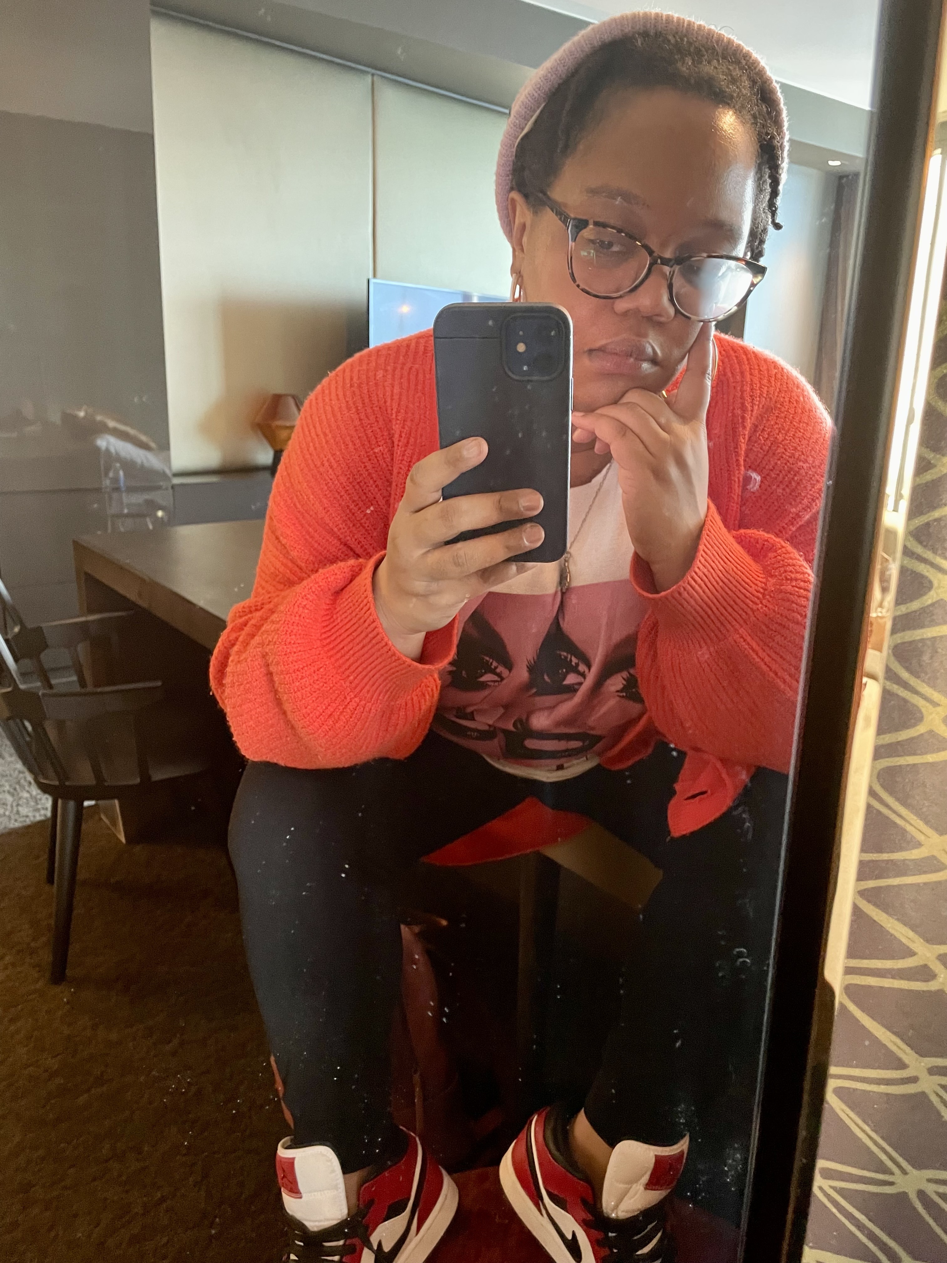 Carmen, a black femme, has a bright orange sweater and a purple beanie, black sweatpants, and red and black Jordans on her feet, she's leaning into a window and taking a selfie.