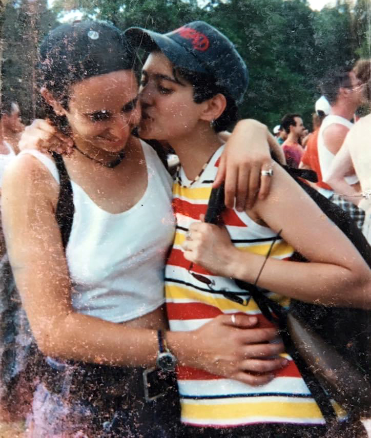 A photo of Tracy and Mia at Lalapalooza. The photo is a bit weathered and worn. Tracy wears a front ways baseball cap, Mia a backwards one. Tracy goes to kiss Mia's cheek and Mia smiles. They both wear tank tops and backpacks.