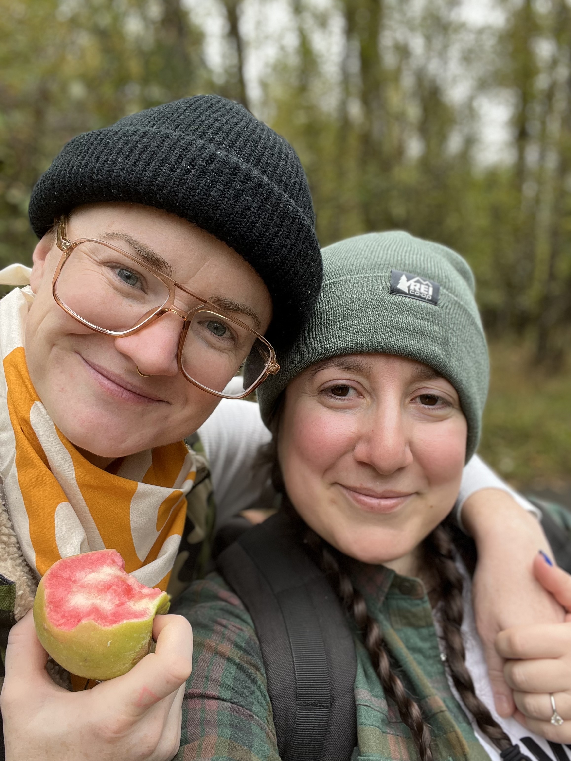 Two dykes take a selfie — one wears an REI bandana and chomps on a green apple with pink insides, the Mountain Rose apple, and the other wears a green REI beanie over her pigtail braids.