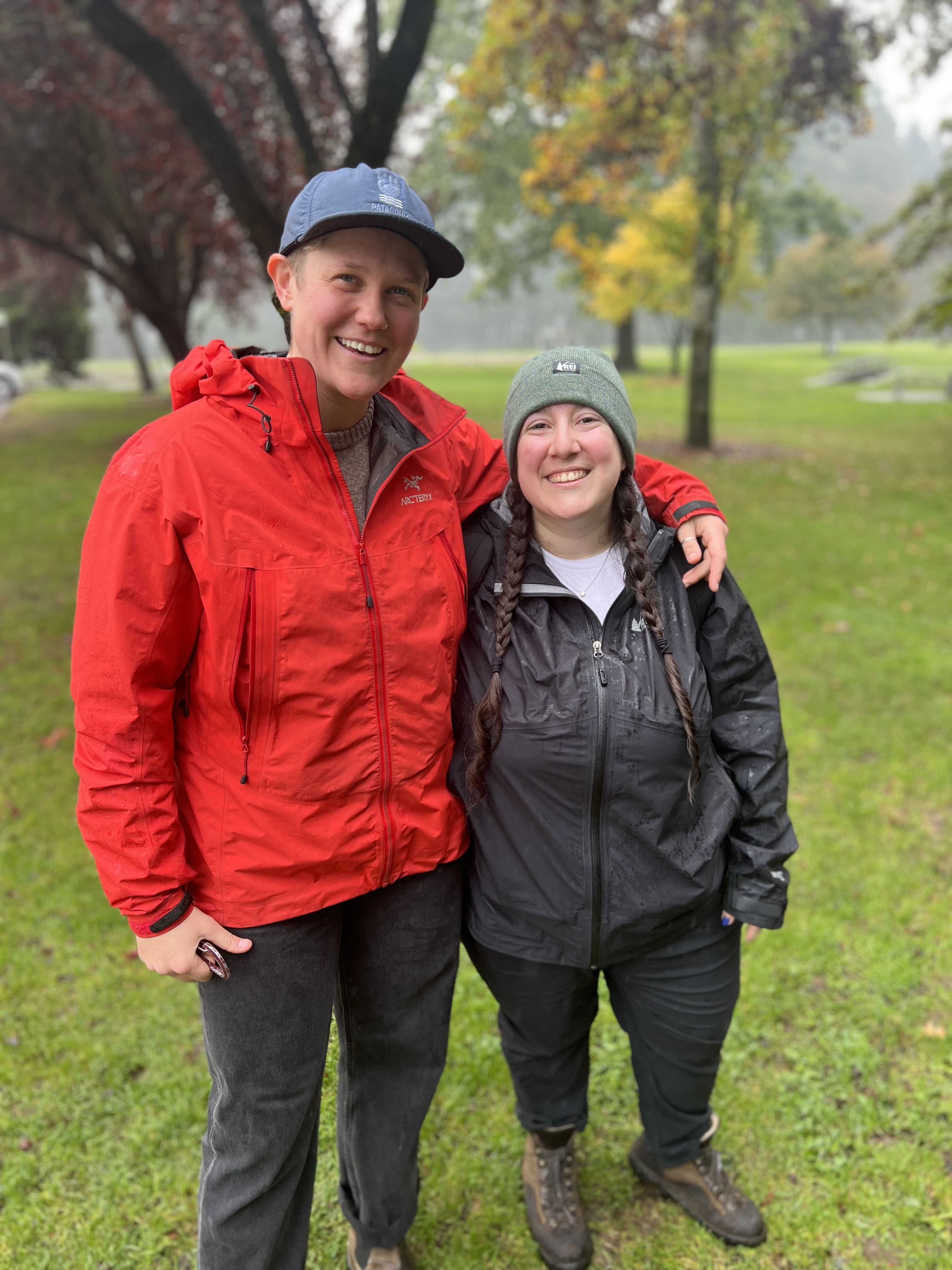 Two queers, Sarah from REI and Vanessa from Autostraddle, pose at the trailhead. Sarah wears a bright red rain jacket and an enormous smile. Vanessa wears a black rain jacket and an equally huge smile.