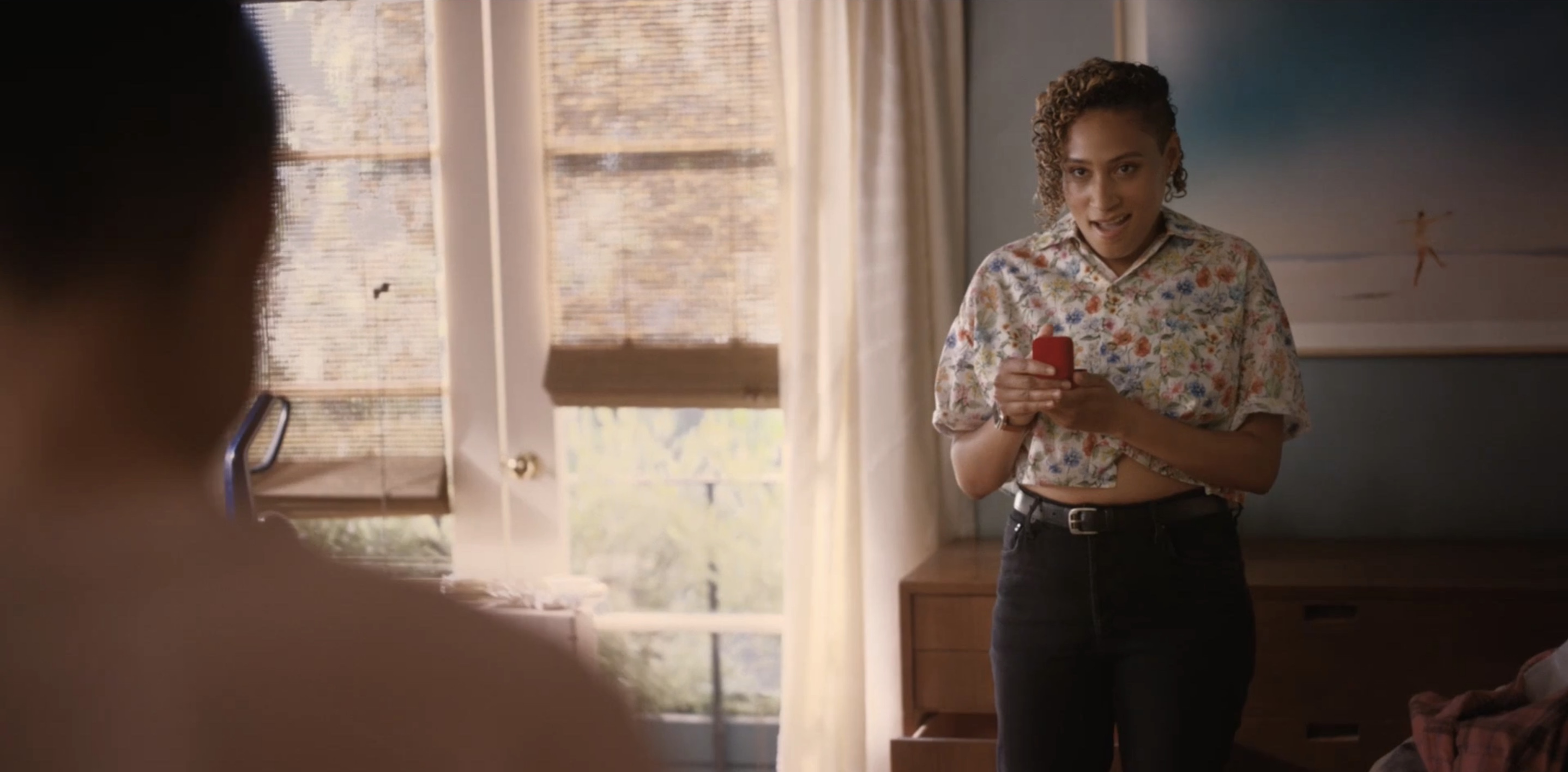 Sophie in a button-up short sleeved floral crop top and black jeans, holding an open jewelry box in amazement