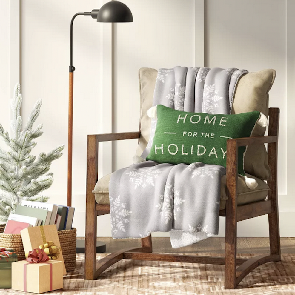 a light gray blanket with white snowflakes draped over a beige and wood chair with a green throw pillow over it. there is a tall brown and black lamp, presents on the floor, and a small pine tree in the corner