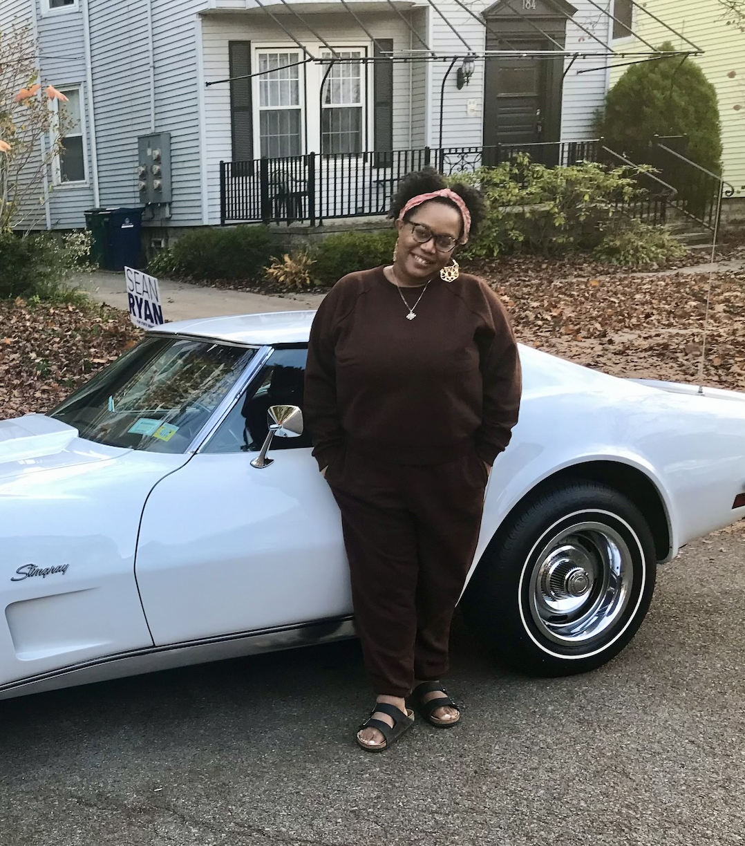 Carmen wears a deep brown sweatsuit and birkenstocks and leans against a car, smiling. Carmen is a Black woman with short black hair and glasses
