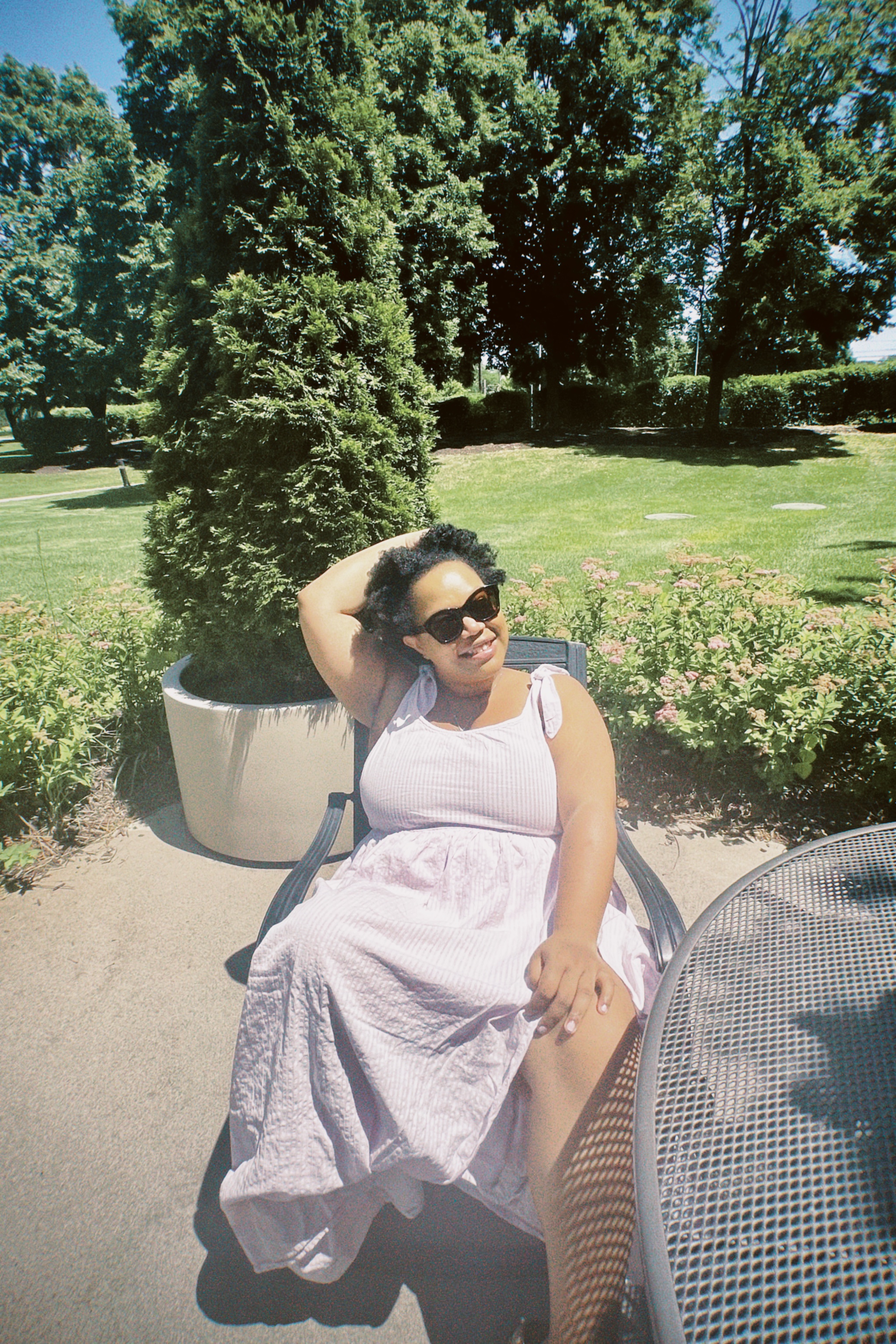 Carmen, a Black woman with glasses and natural hair, is sitting in a chair in a park in a light purple dress and sunglasses