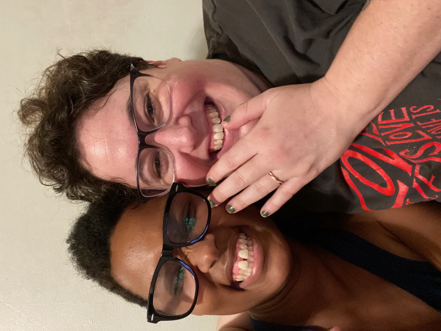 Sai, a black woman with short hair is wearing black glasses and smiling. Beth, a white woman with short hair is wearing glasses and smiling. She is holding up her left hand, a small rose gold band is on her ring finger