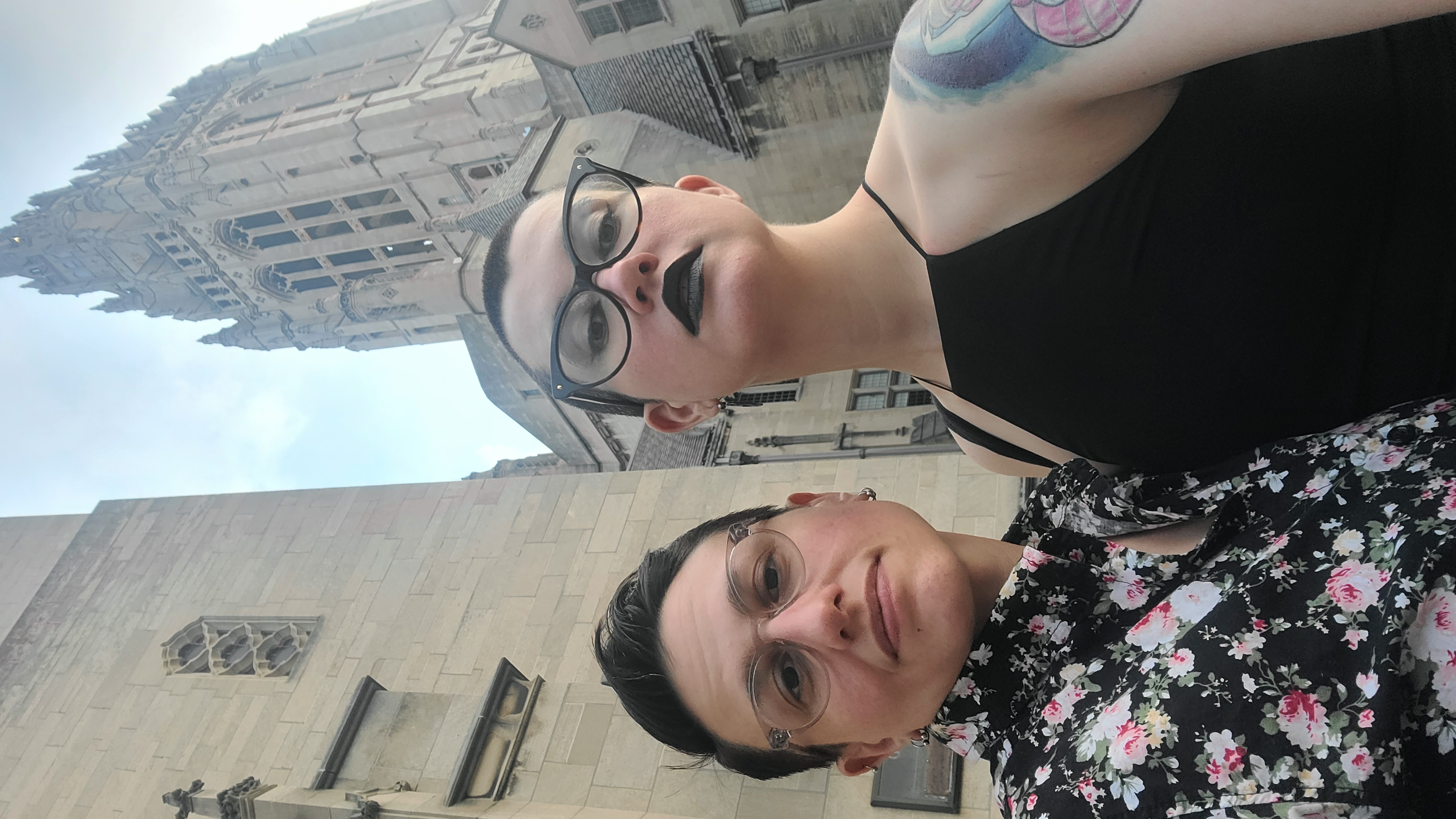 nico and sadie are dressed up and in front of a church. nico has a shaved head and is wearing a black dress and black lipstick. sadie is wearing a floral print button down shirt. they both wear glasses