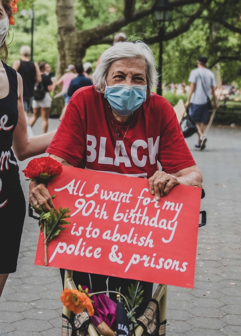 Shatzi Weisberger, known as The People's Bubbie, at another one of the 2020 Black Lives Matter uprisings. This time she wears a red t-shirt, a blue mask, and holds a red rose and a red sign with white lettering that says All I Want For My 90th Birthday Is To Abolish Police And Prisons