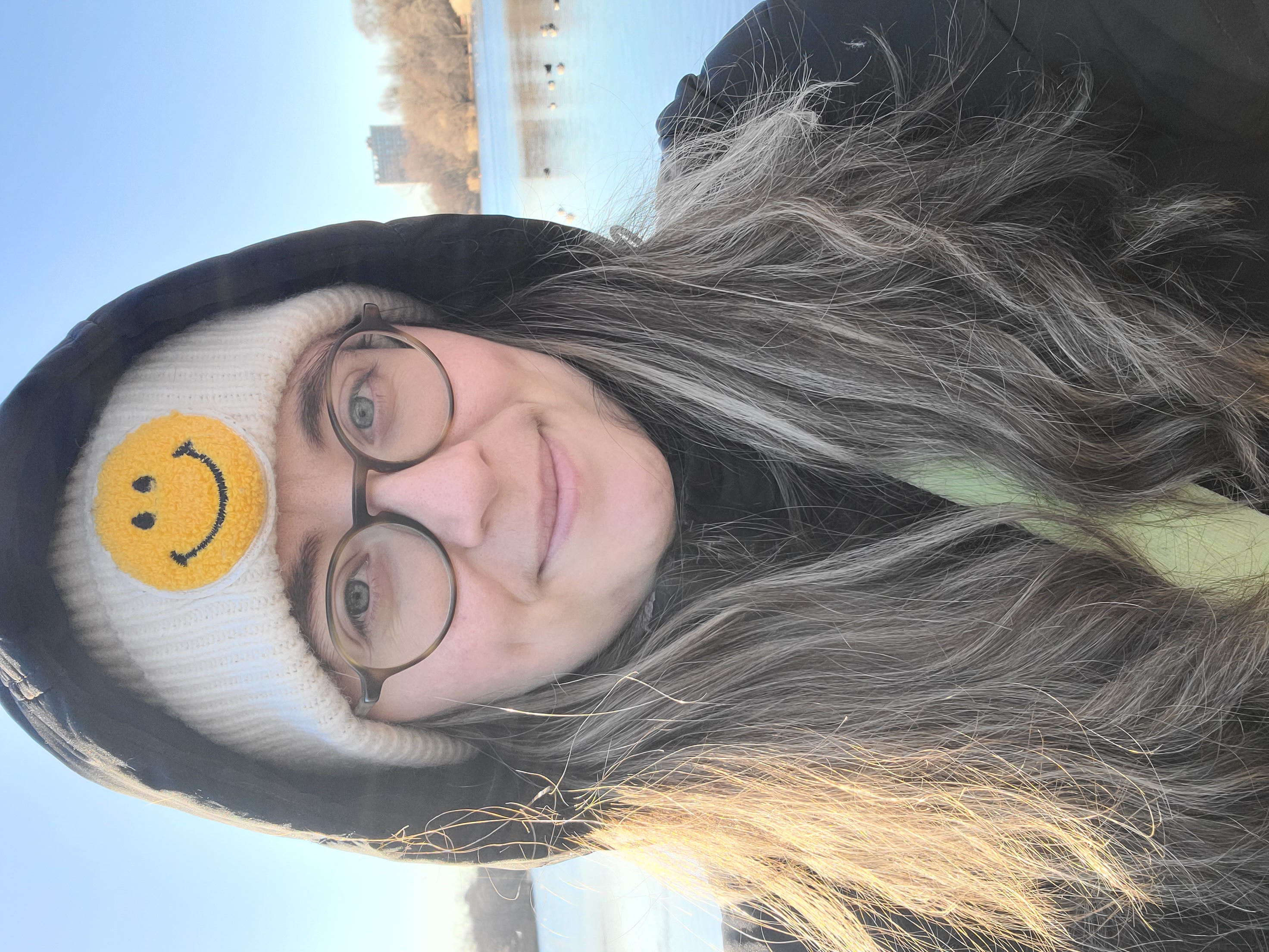 anya, a white woman with glasses and long brown hair, smiles while wearing a beanie with a smilie face on it and a jacket with a hood in front of a lake
