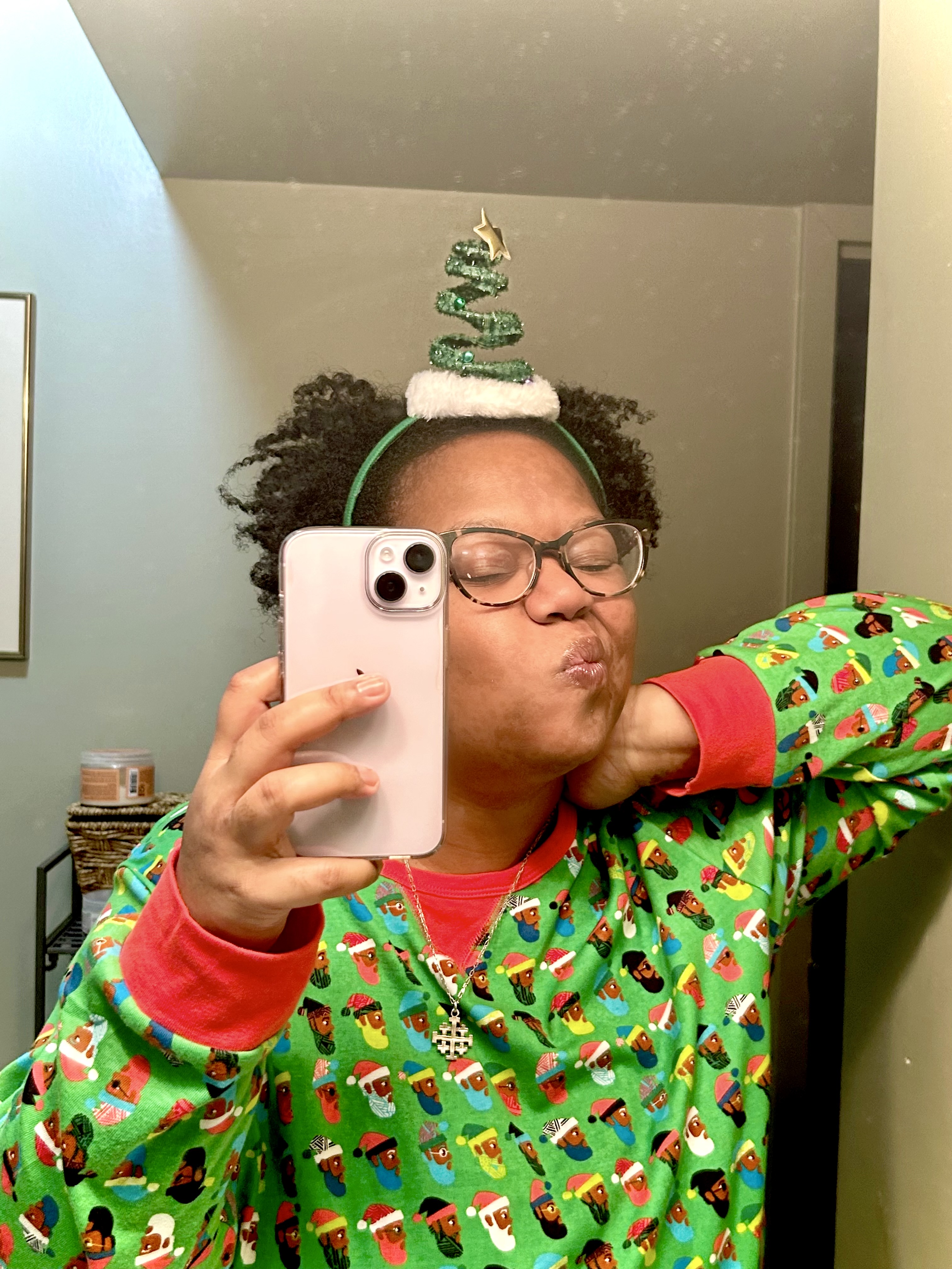carmen a black woman with short curly hair and glasses makes a silly face while wearing red and green santa face patterned pyjamas and a christmas tree headband