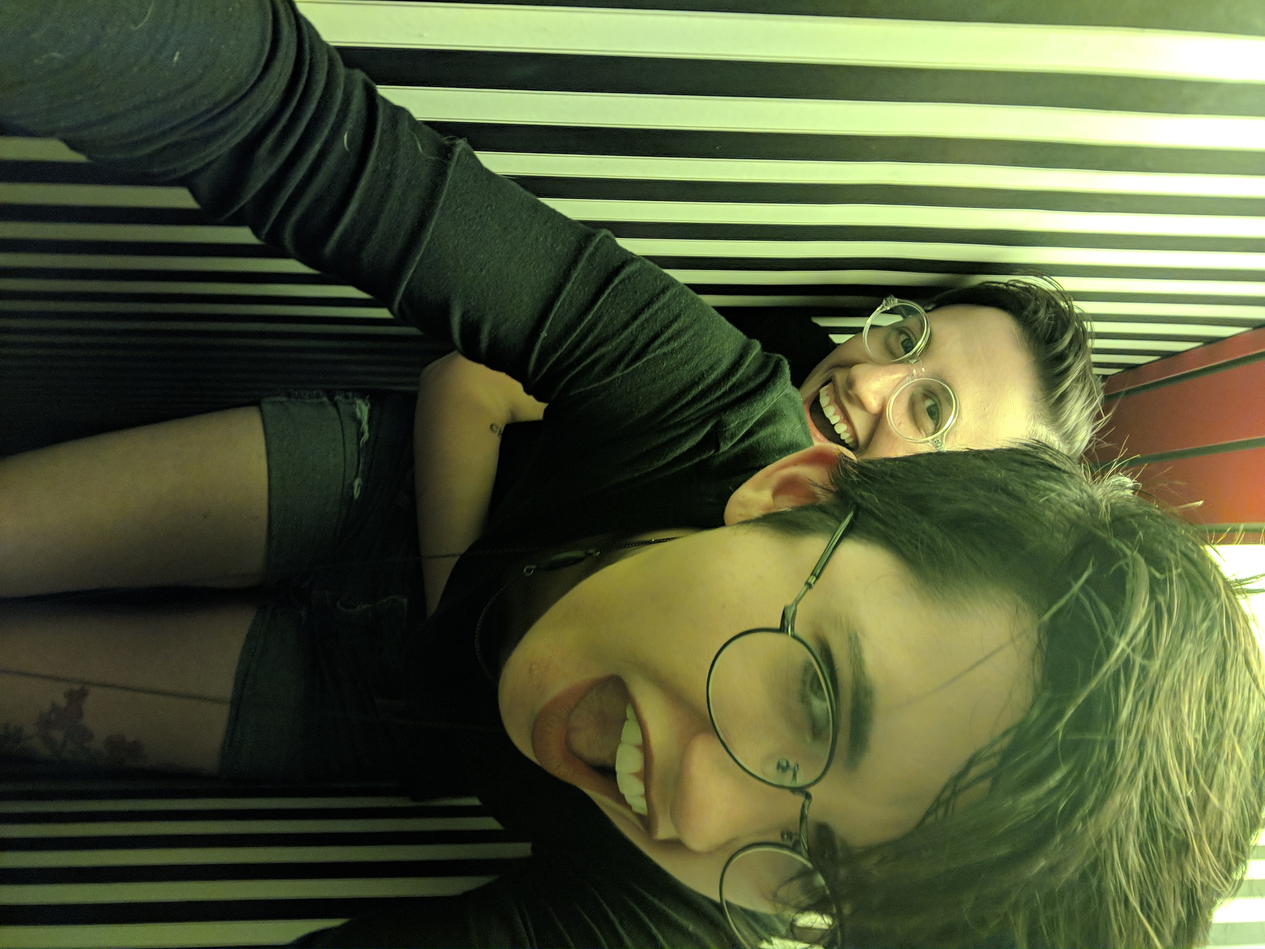 Nico and Sadie are in a greenlit striped hallway that is very narrow inside of an art house. Sadie grabs Nico from behind and Nico makes a mock affronted face.