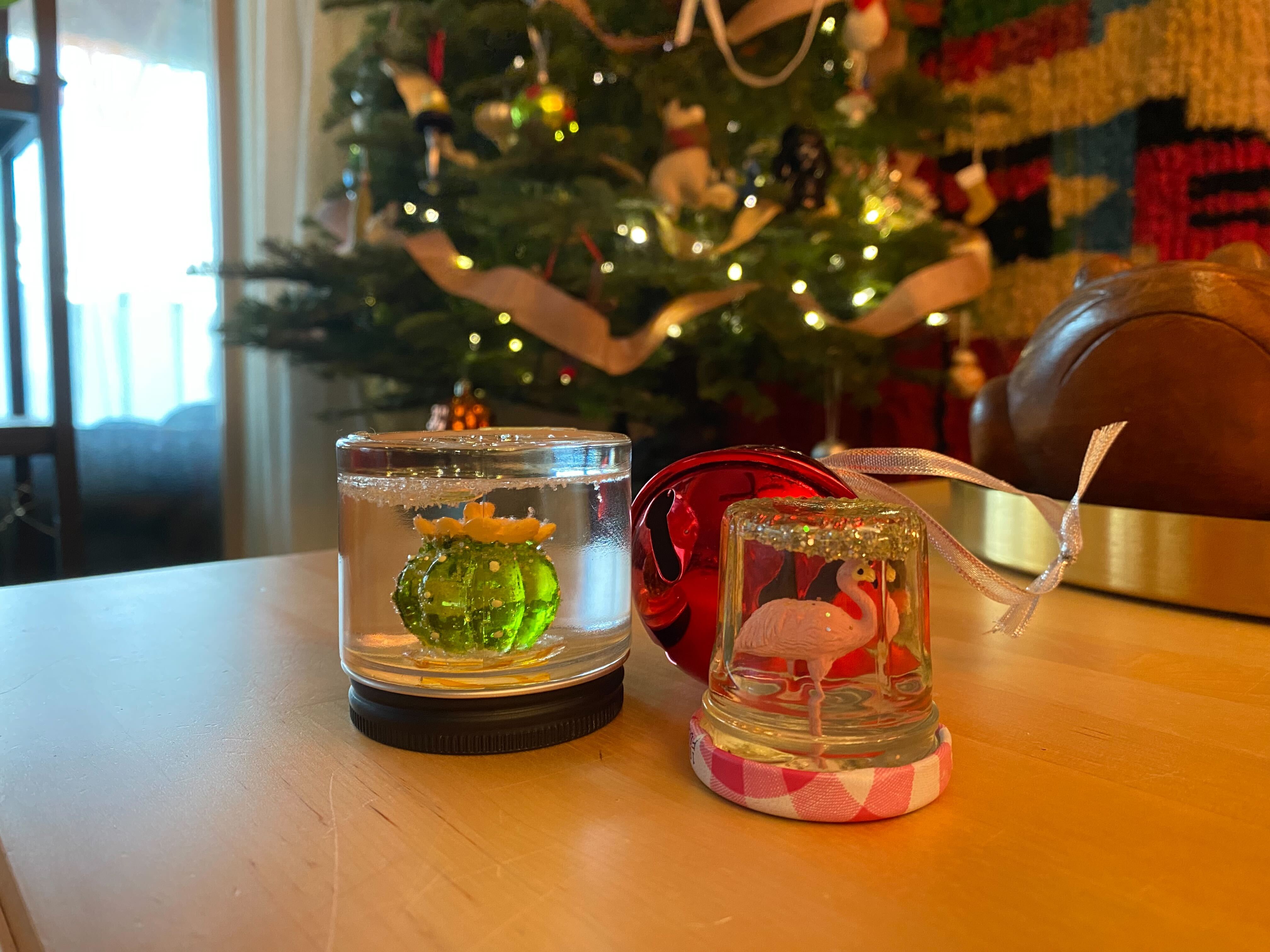 Laneia has posed two completed snow globes in front of her Christmas tree. One contains a flamingo figurine and the other a cactus figurine. They are sparkly and the mood is warm and festive.