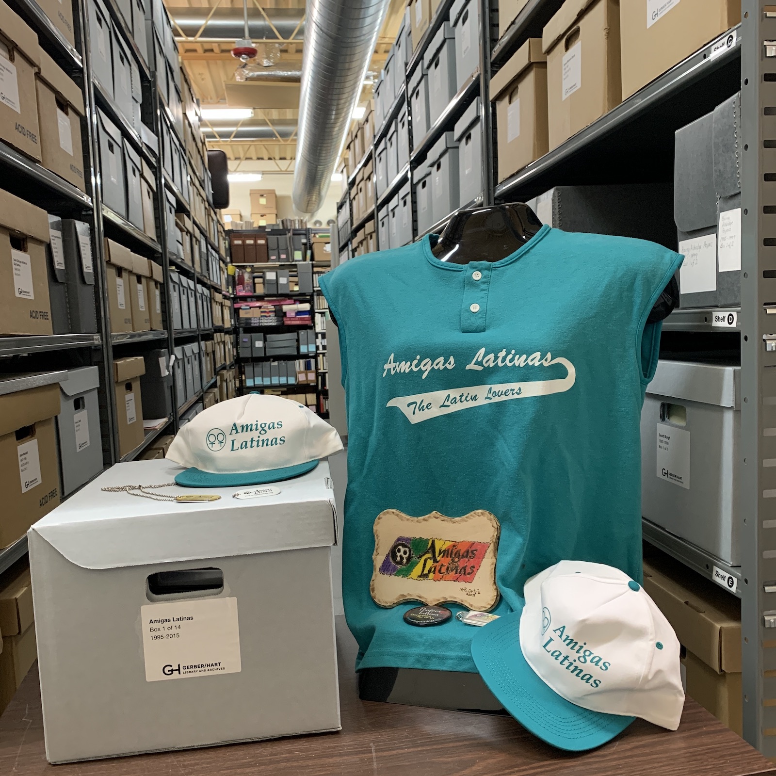 a photo of the Gerber/Hart Archives, featuring many shelves with many boxes in the background, and in the foreground materials from the Amigas Latinas collection: a bright blue tank top and a blue and white hat with the words Amigas Latinas on them, next to a closed box of other materials
