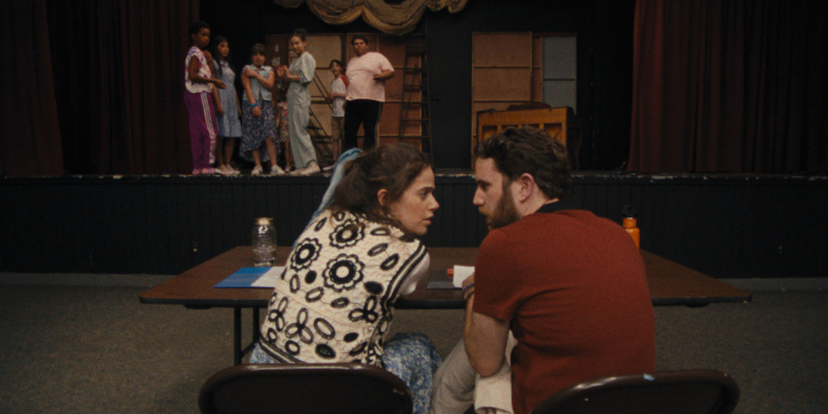 Molly Gordon and Ben Platt sit together at a directors table in a theater. Several people are on stage looking down at them.