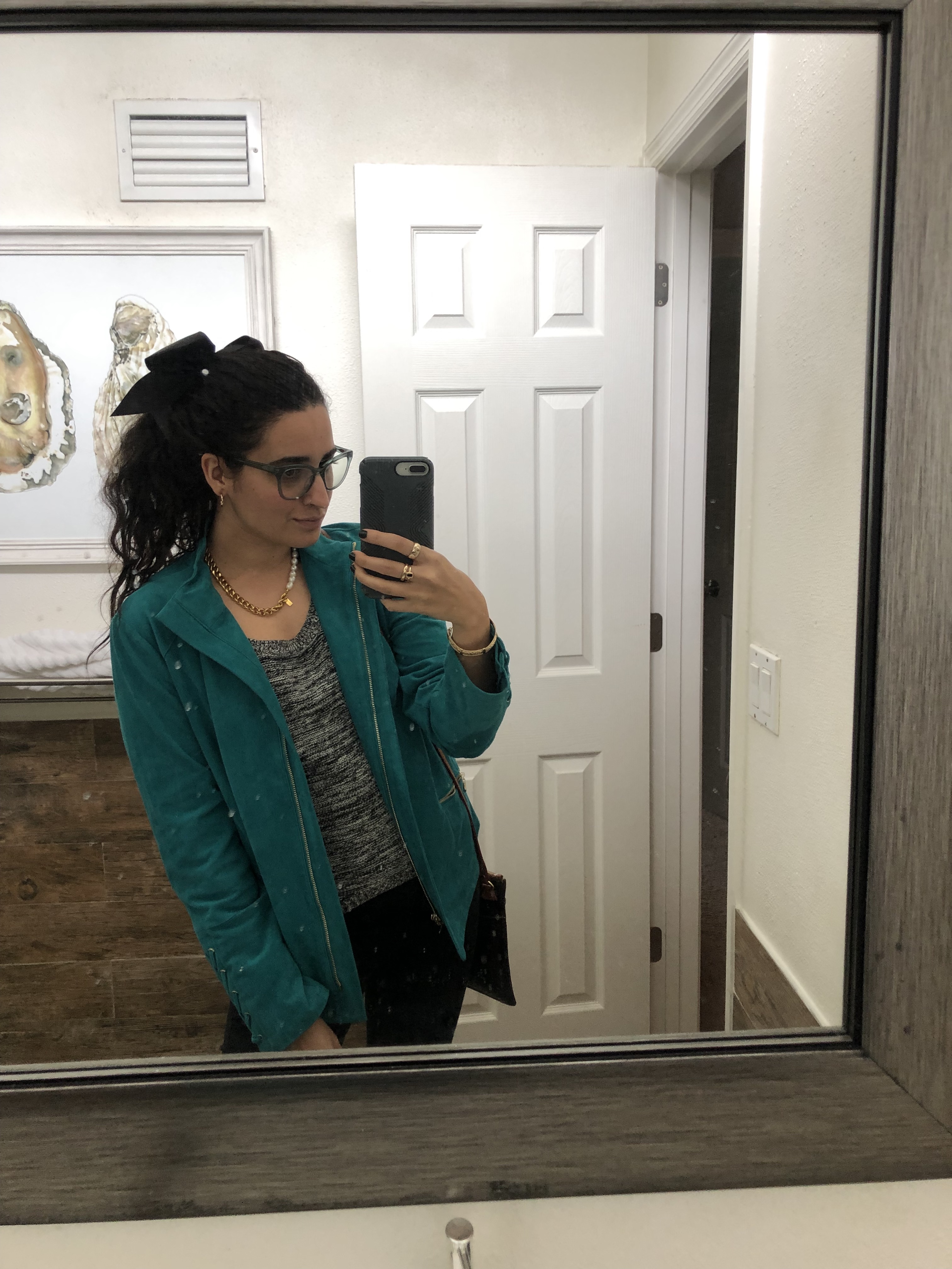 Kayla Kumari Upadhyaya wears a black and white knit blend sweater and black jeans and a teal suede motojacket. Her hair is in a ponytail with a black bow and she wears blue framed glasses and a chunky gold chain and pearl necklace.