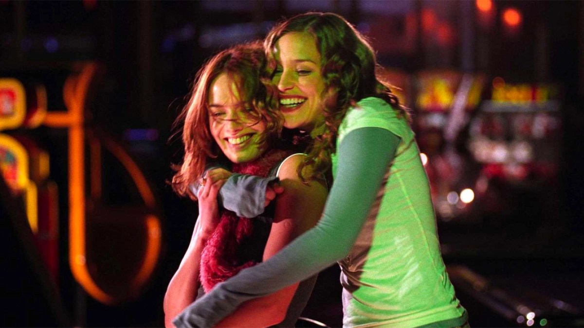 In a still from the movie Imagine Me & You, two white lesbians hold each other in a hug while laughing. They are wearing classic 00s fashion, including a useless scarf and a short sleeved t-shirt over a long sleeved t-shirt.