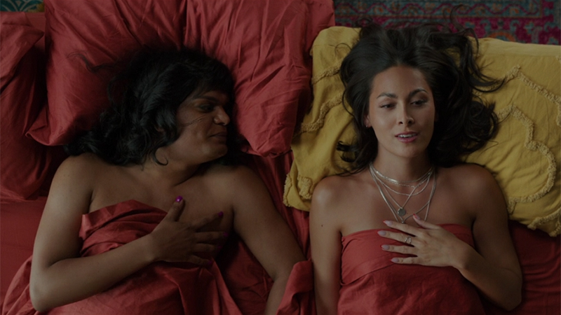 Two trans femmes, Sabi and Olympia are in a bed made with blood orange sheets. Sabi looks at Olympia, who's lying on a mustard yellow pillow.
