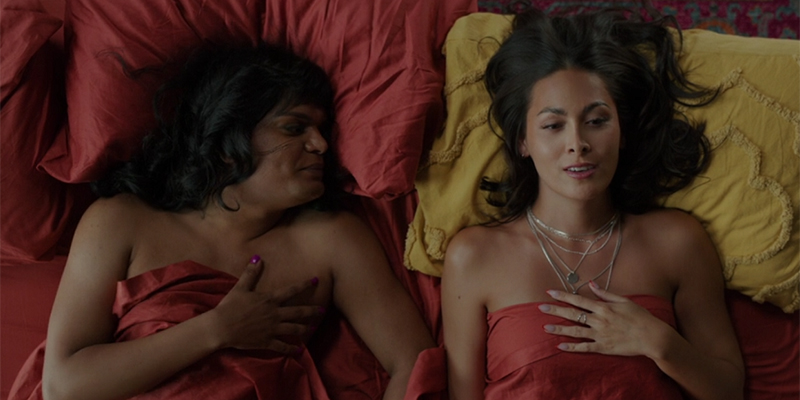 Two trans femmes, Sabi and Olympia are in a bed made with blood orange sheets. Sabi looks at Olympia, who's lying on a mustard yellow pillow.