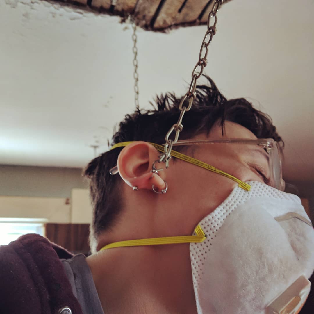 a close up of sadie, a butch woman wearing an n-95 mask, who has her earring caught on a drop ceiling chain